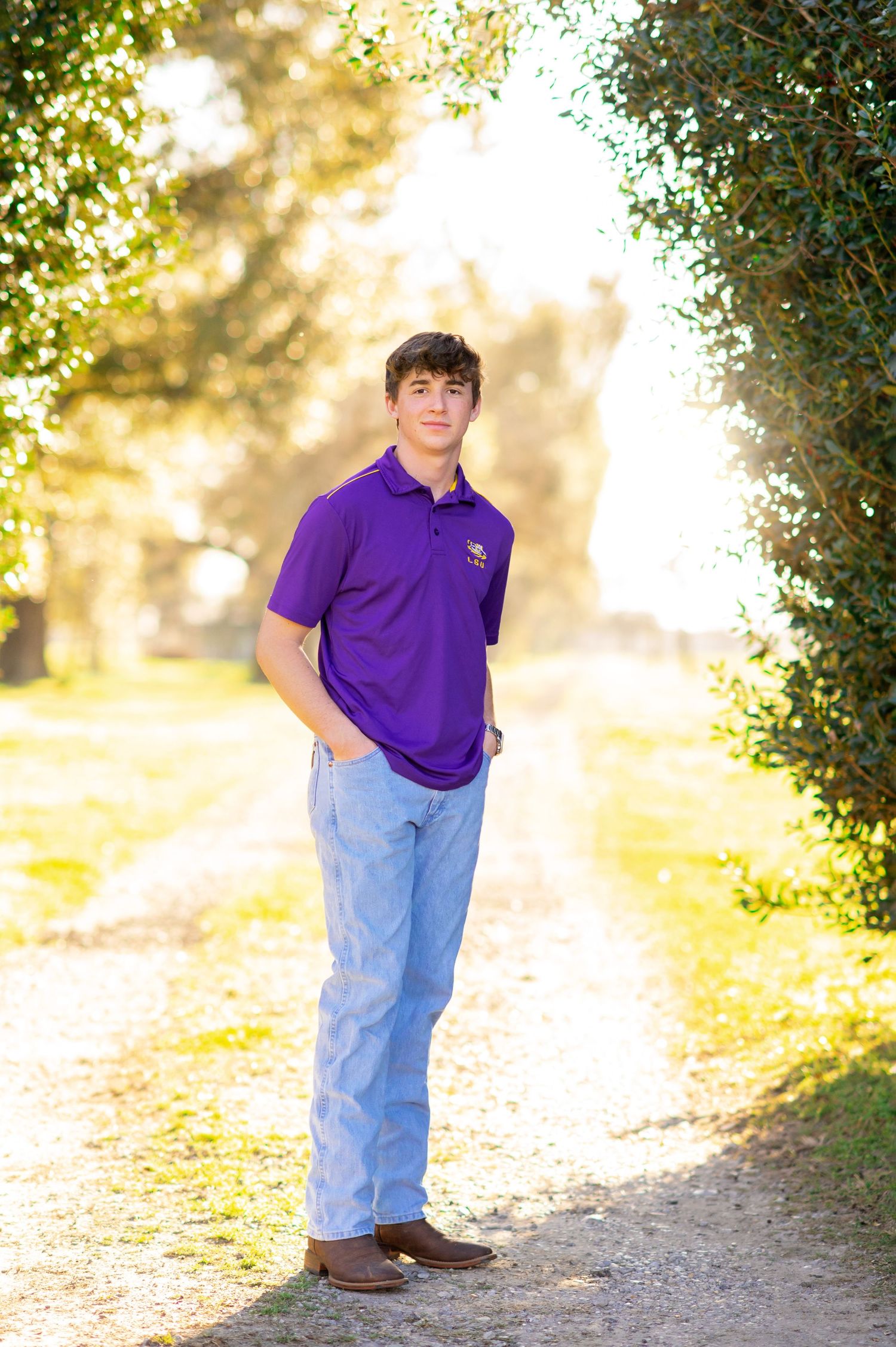 Young man in purple polo shirt and jeans posing on a sunlit tree-lined path for senior portraits.