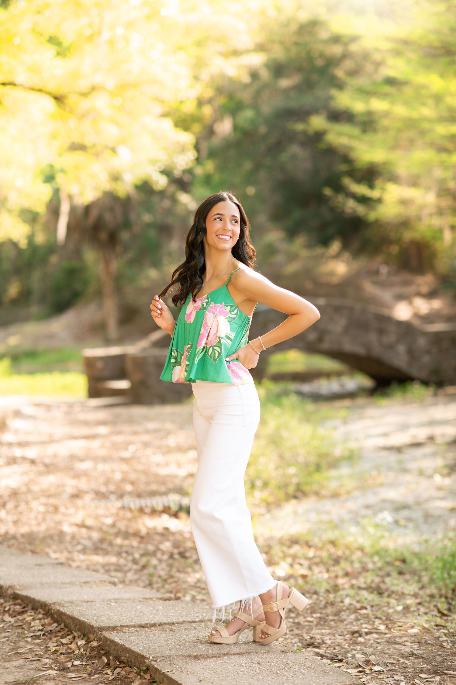 Young woman in green floral top and white wide-leg pants posing outdoors in a sunlit park with stone bridge.
