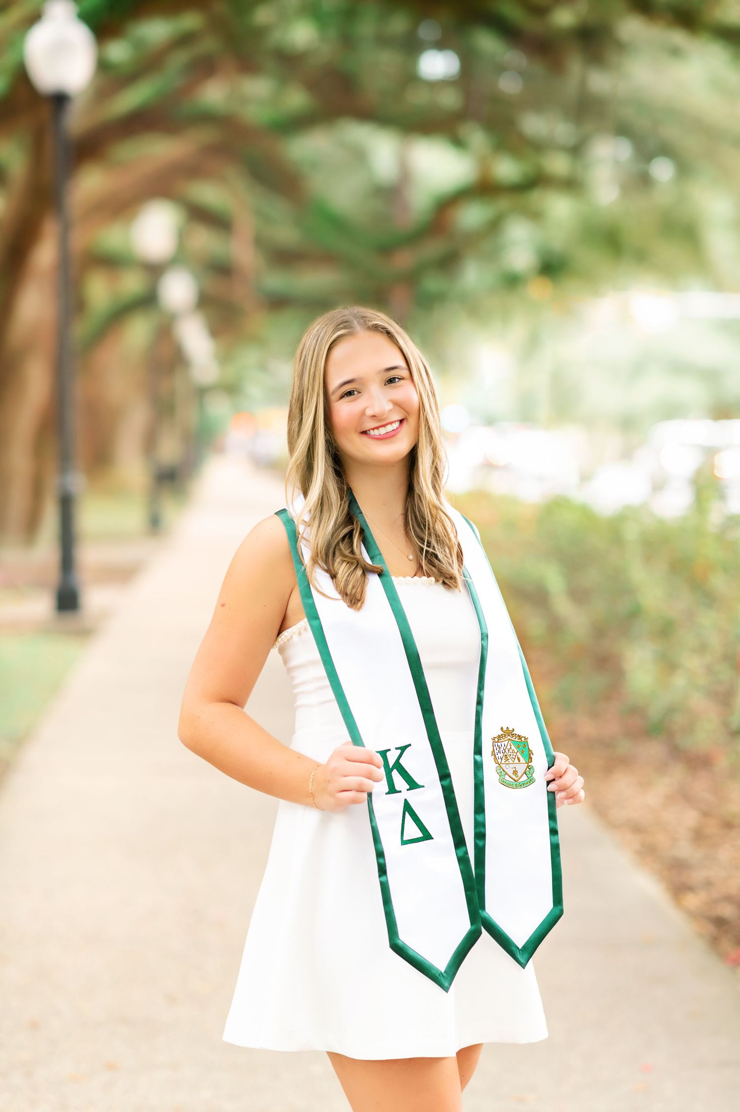 Smiling graduate in white dress wearing Kappa Delta sorority stole on a tree-lined campus walkway.