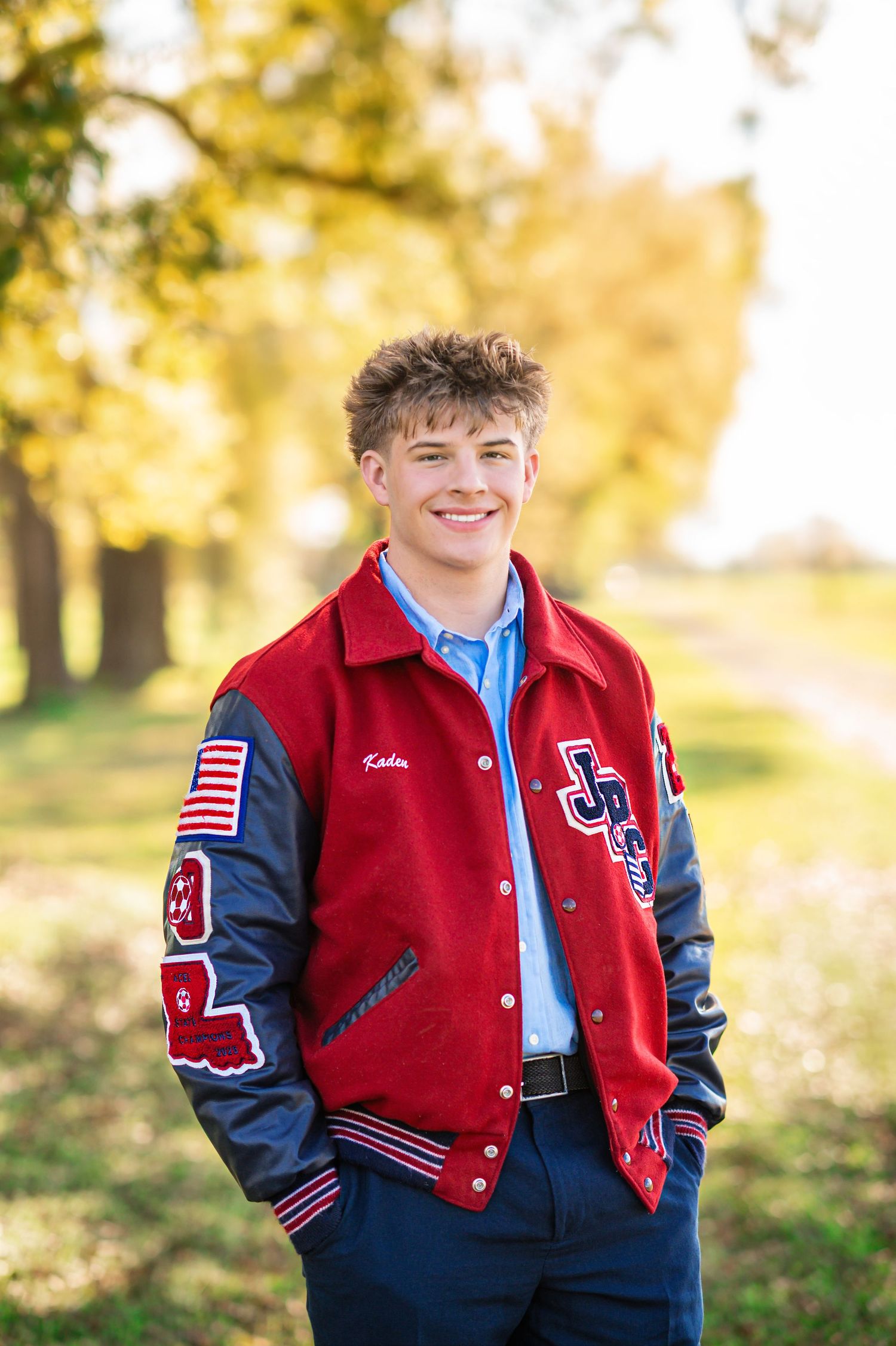 Smiling teen boy in red varsity letterman jacket poses outdoors among fall foliage for senior portrait.