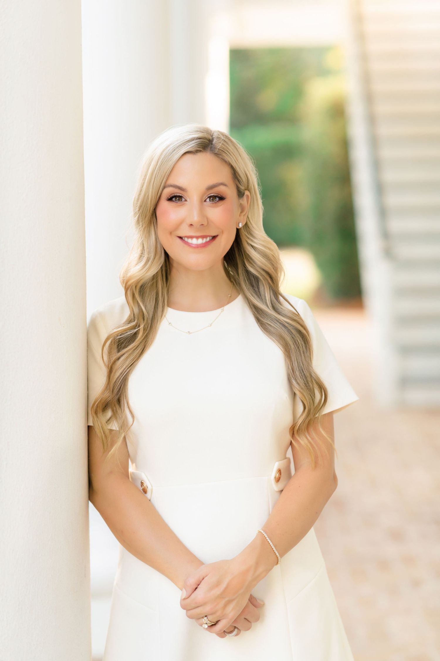 Smiling blonde woman in white dress leaning against white column with soft bokeh background.