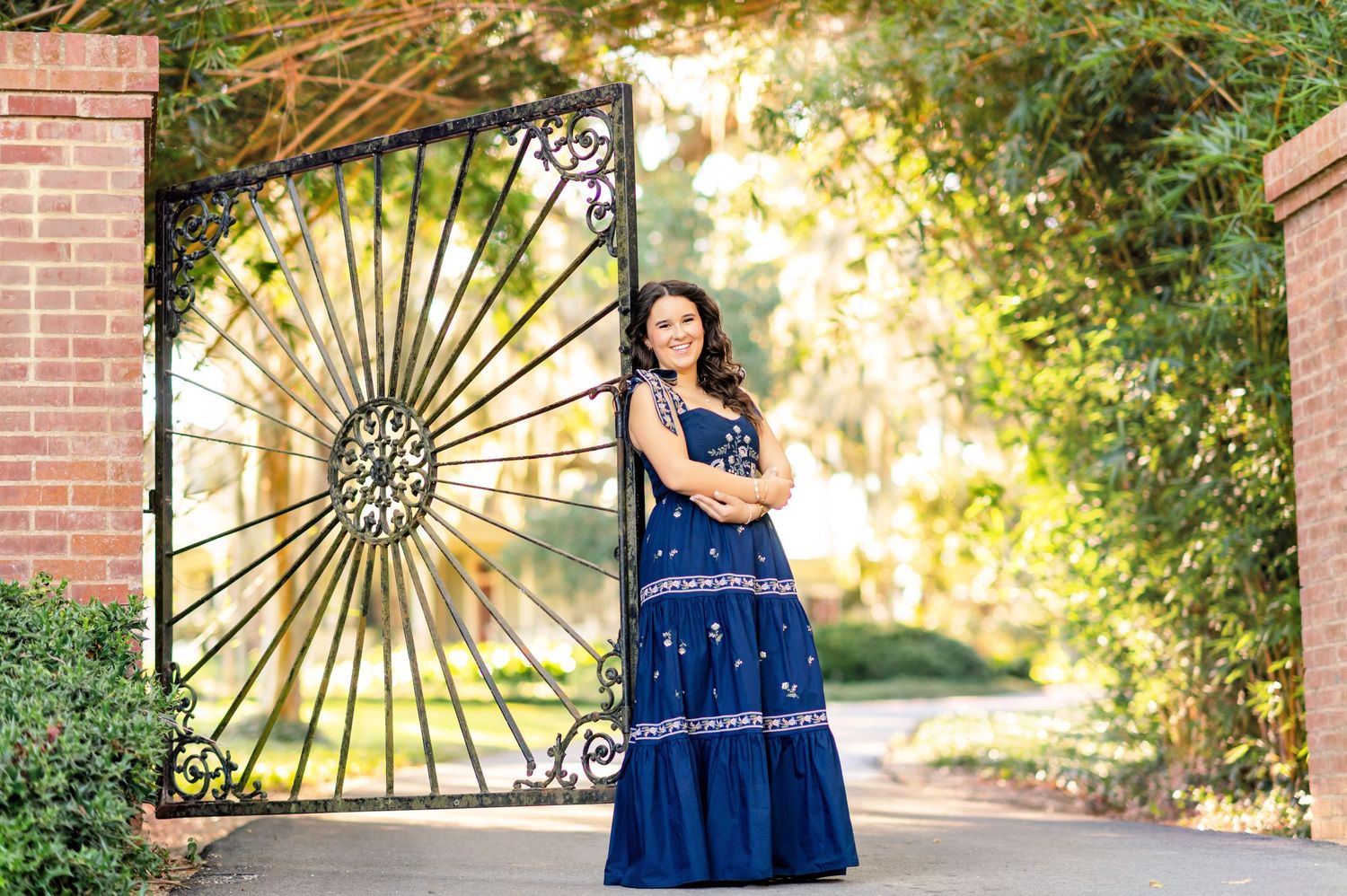 Woman in blue embroidered maxi dress posing by ornate iron gate with brick pillars and lush greenery.