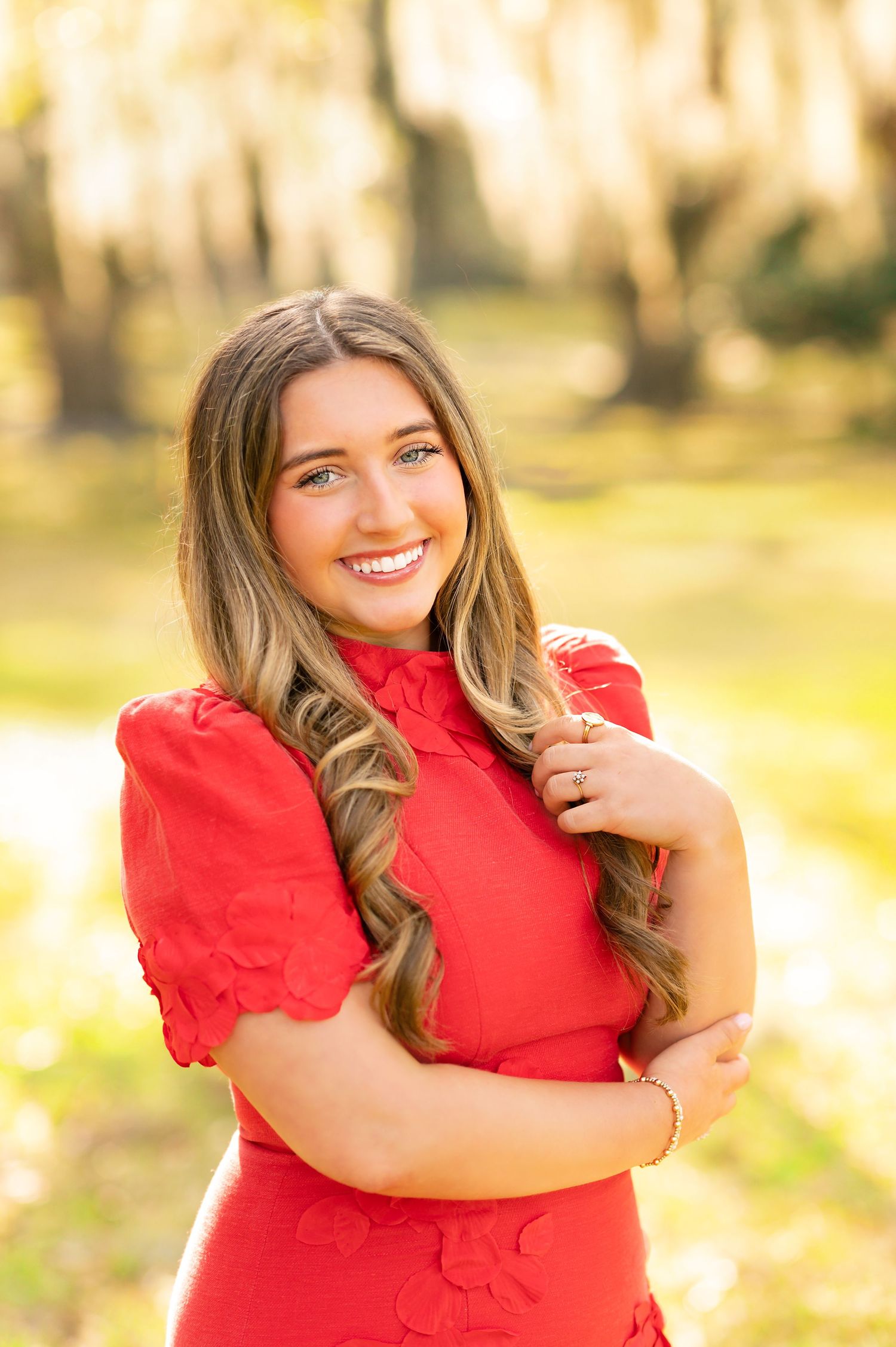 Young woman in red dress with long wavy hair smiling outdoors in golden sunlight among trees.