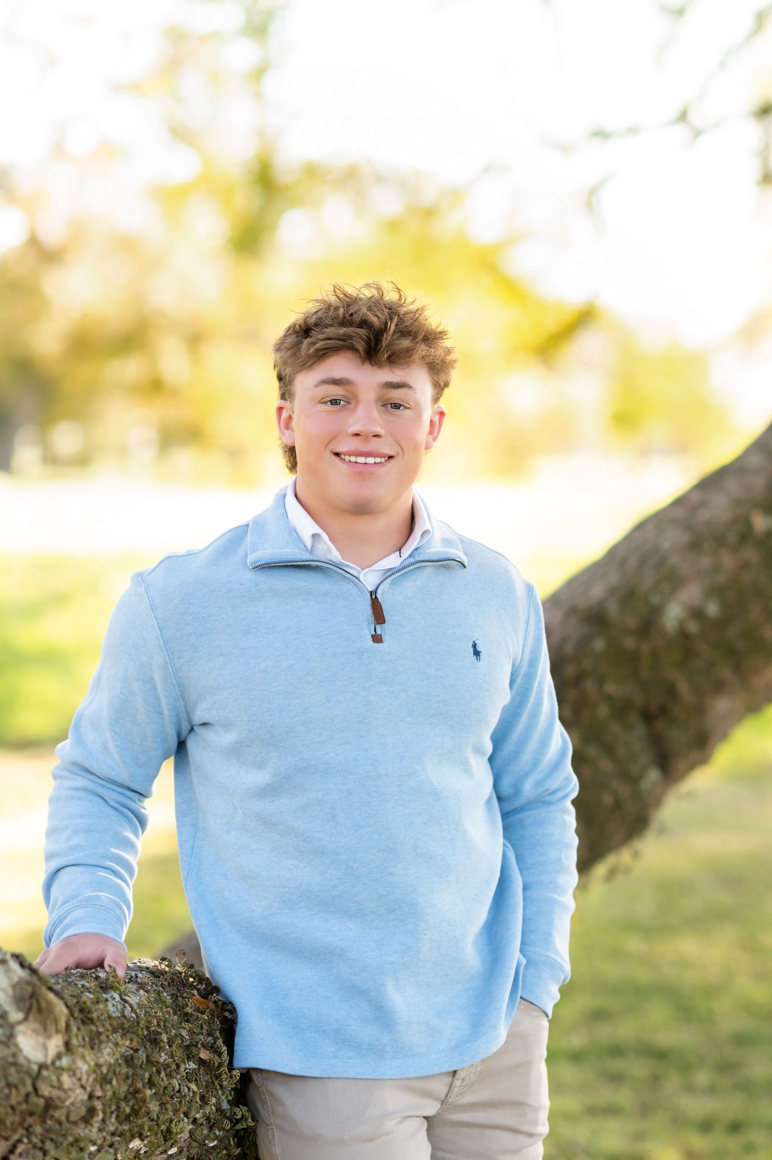 Smiling teenage boy in light blue Ralph Lauren quarter-zip sweater leaning against a tree outdoors.