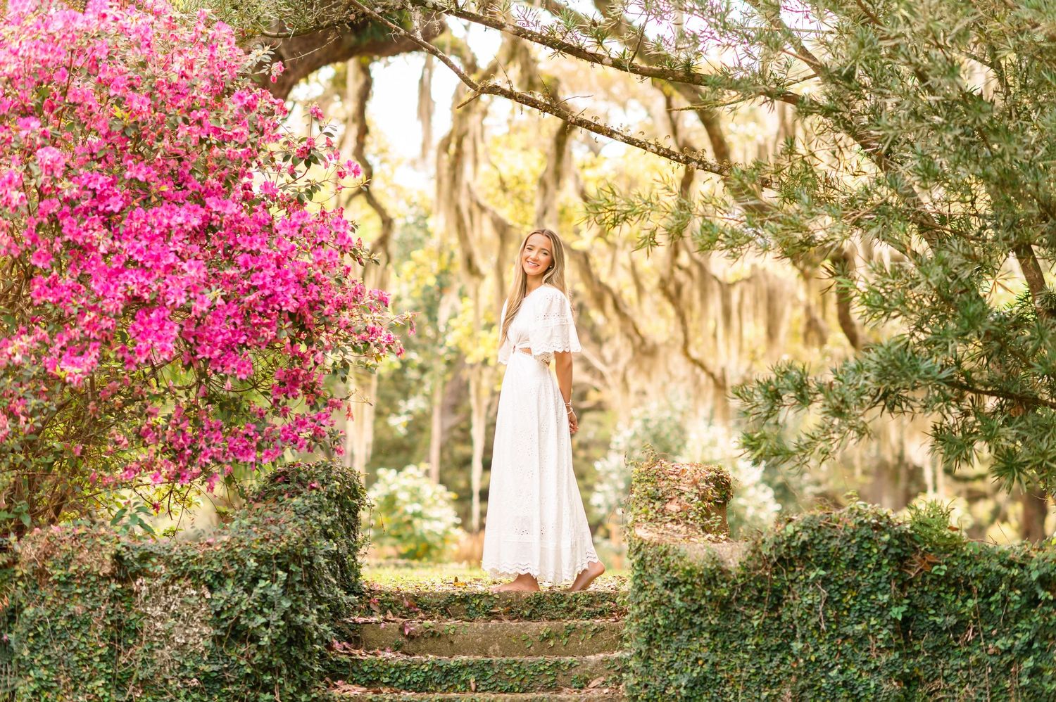 Woman in white dress poses on stone steps surrounded by pink azaleas and moss-draped oak trees in a lush garden.