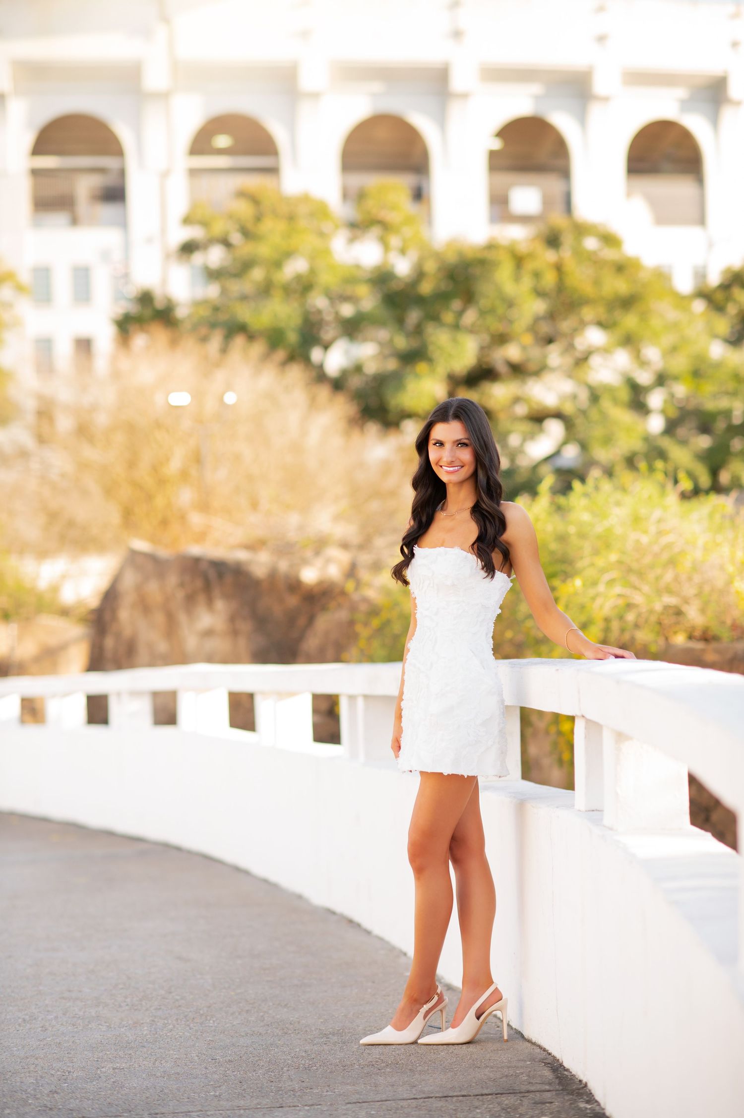 Elegant woman in white strapless mini dress posing on white bridge with classical building backdrop.
