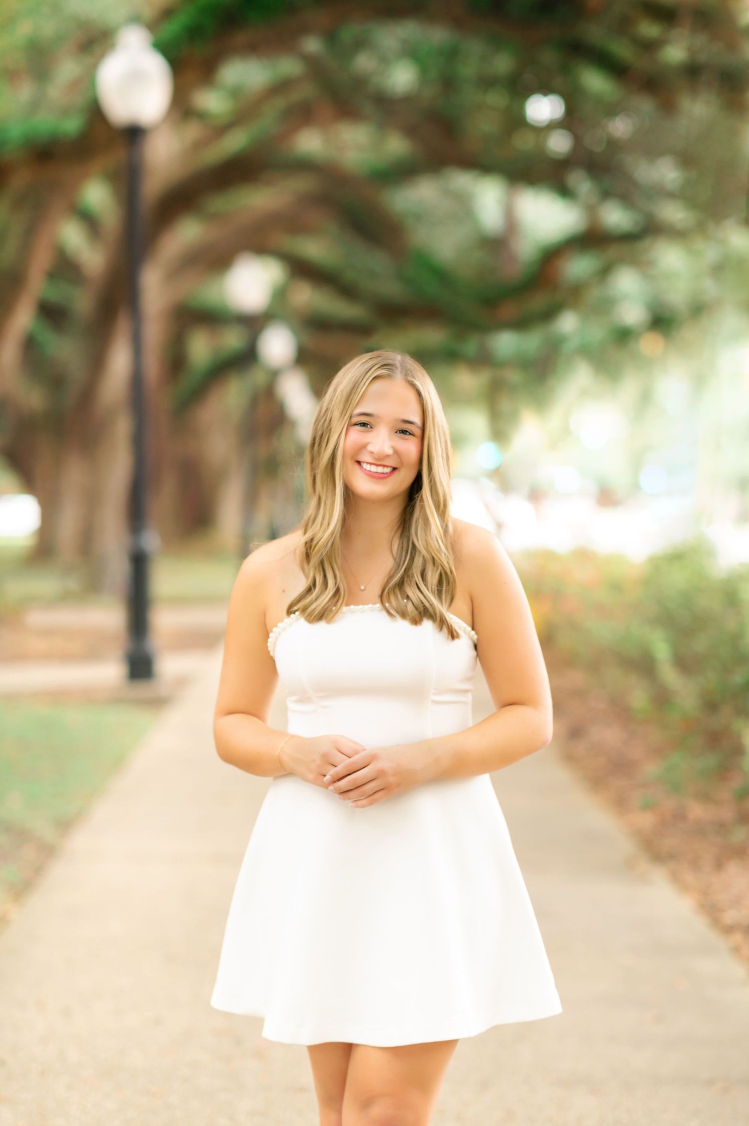 Young woman in white strapless dress smiling on tree-lined sidewalk path during outdoor senior portrait session.
