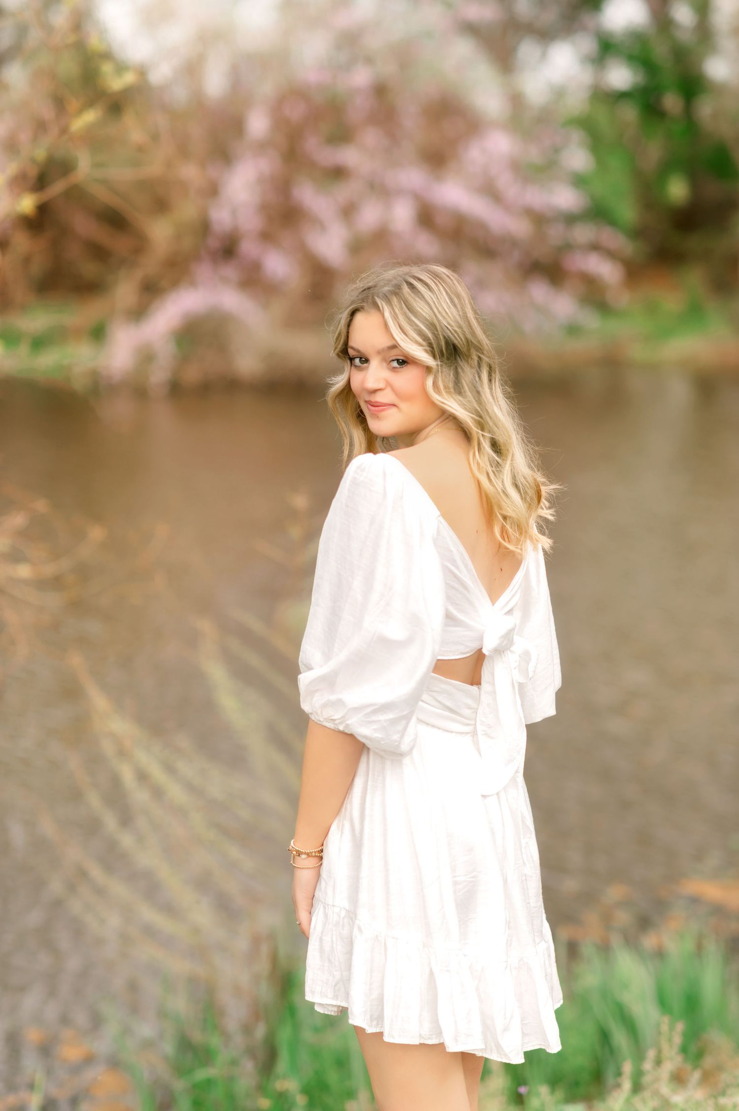 Blonde woman in white backless dress poses near a lake with blooming trees in soft natural light.