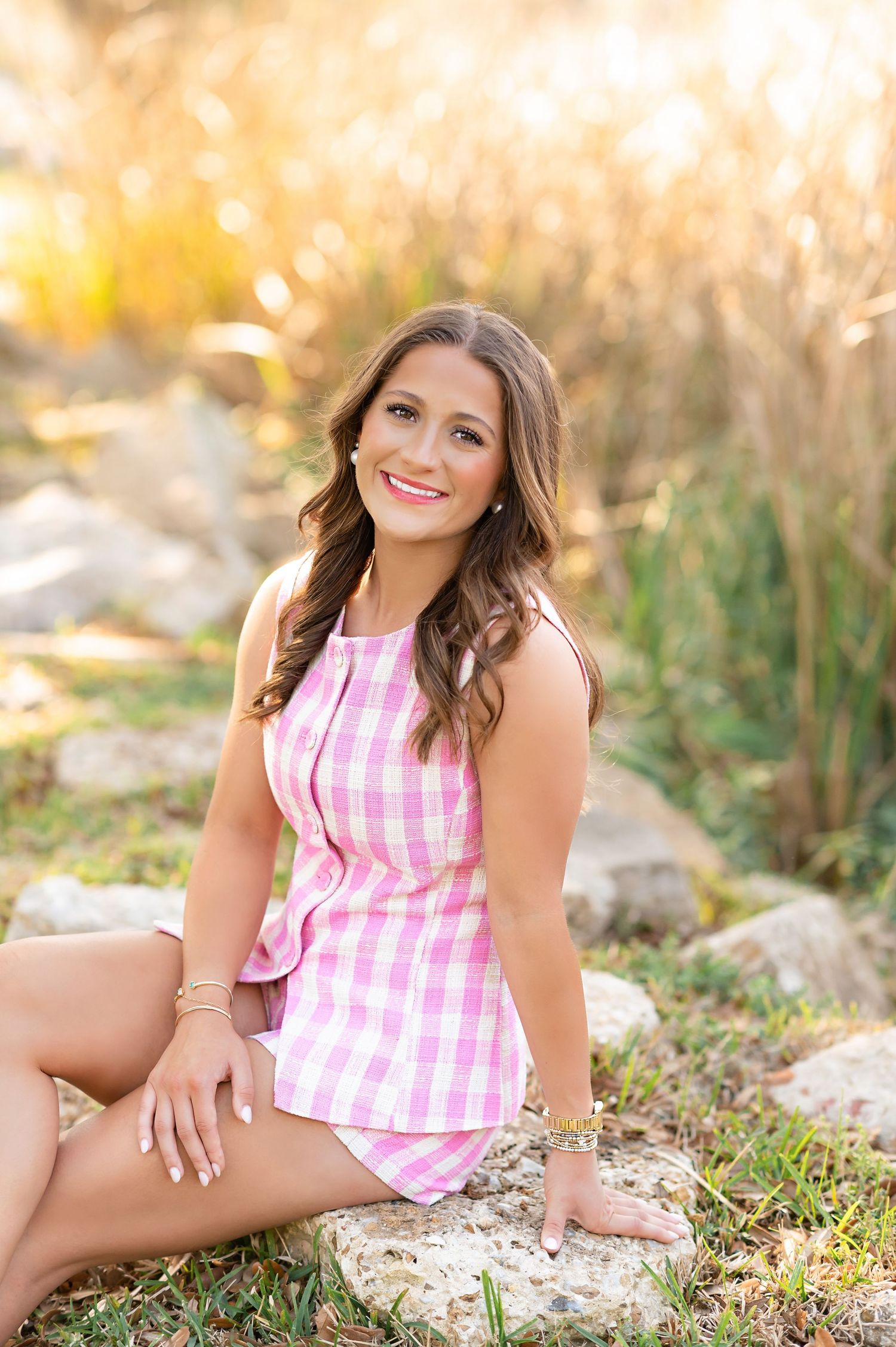 Young woman in pink gingham outfit sitting on rocks outdoors, smiling in golden sunlight.