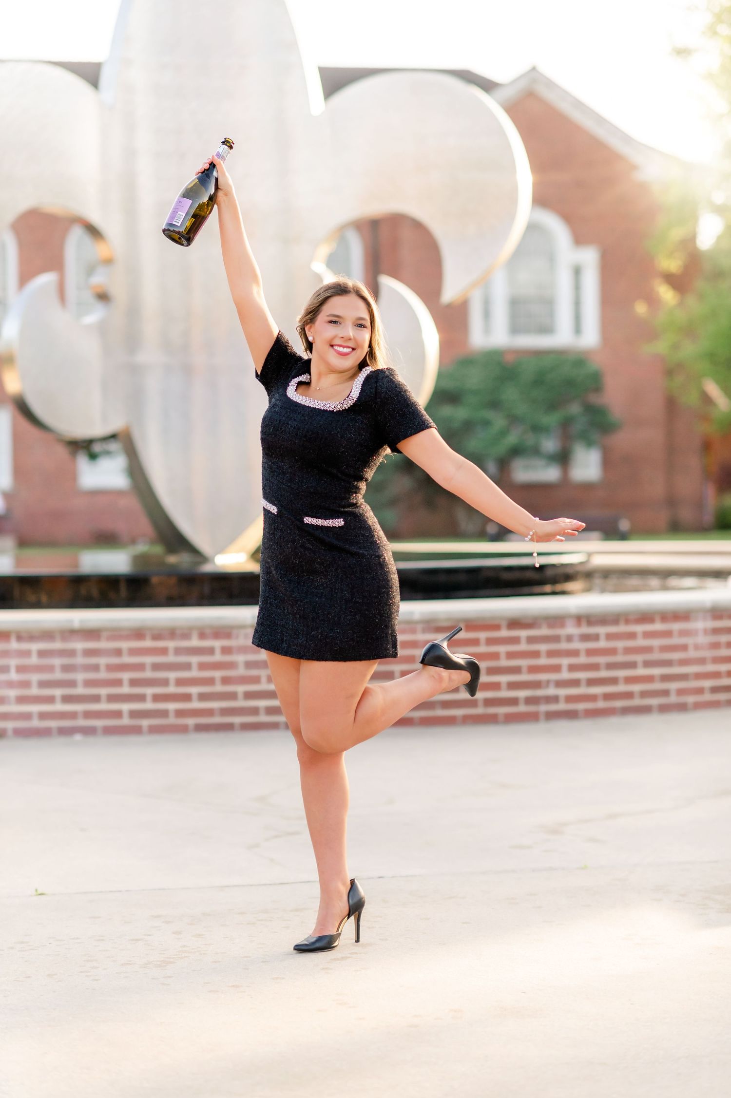 Joyful young woman in black dress celebrates holding champagne bottle aloft near fleur-de-lis sculpture.