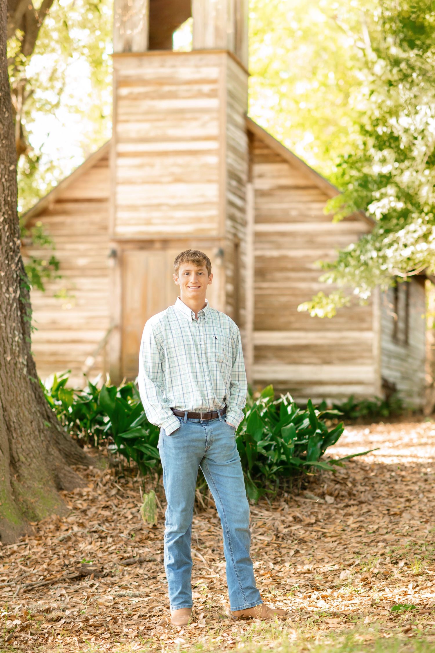 Young man in plaid shirt and jeans smiling outdoors near rustic wooden structure surrounded by lush greenery.