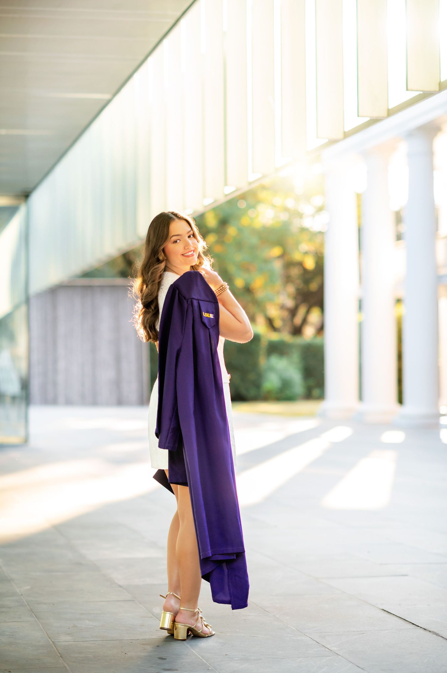 Young woman in purple graduation gown posing outdoors in golden hour light, wearing gold heels.