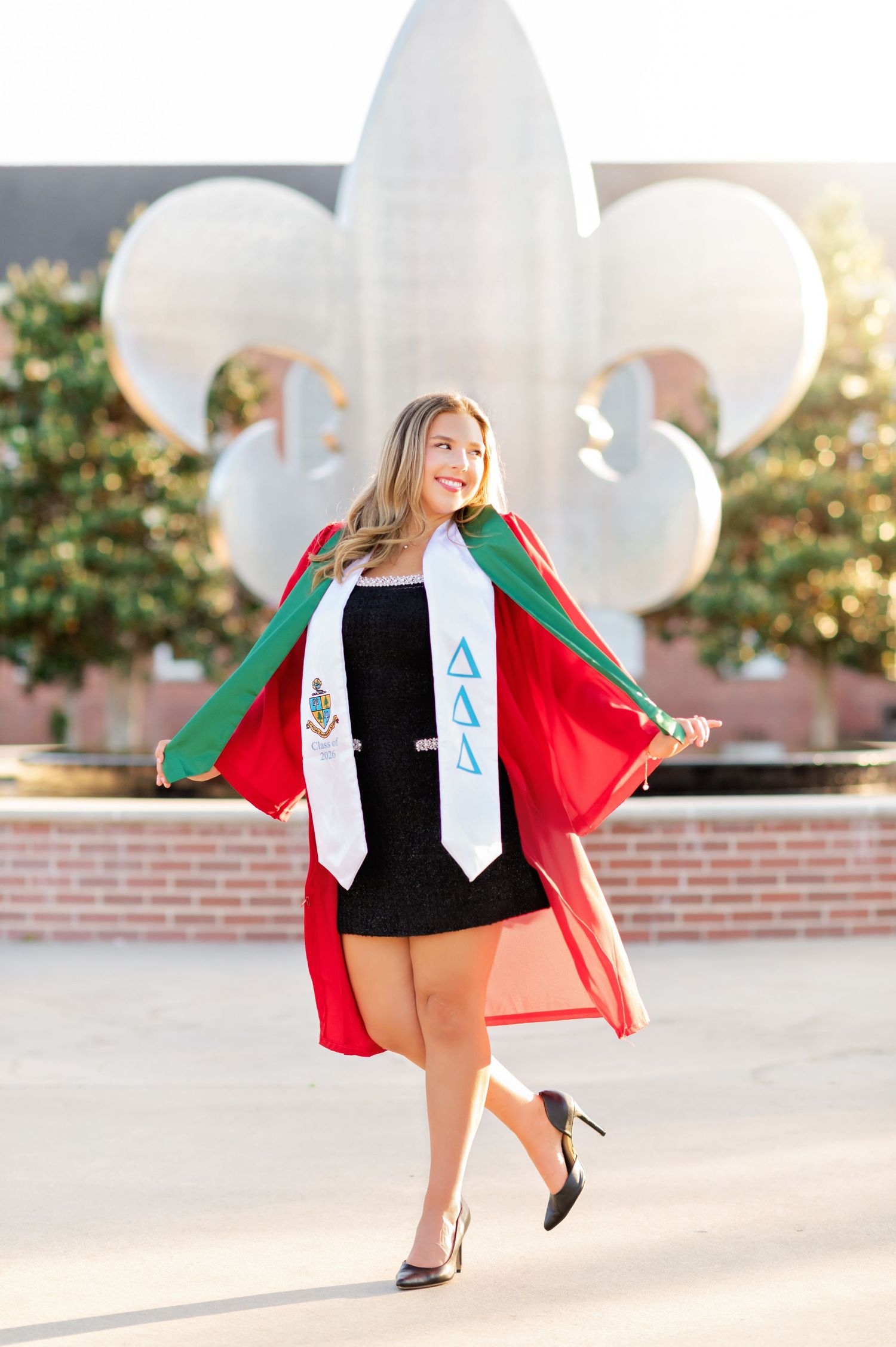 Smiling graduate in a black dress twirling her red graduation gown with Delta Delta Delta stole outdoors.