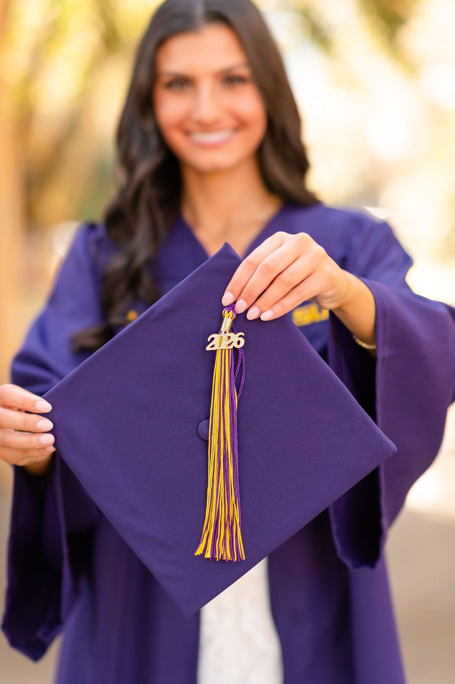 A smiling graduate in purple cap and gown holds up her 2026 graduation cap with gold tassel outdoors.