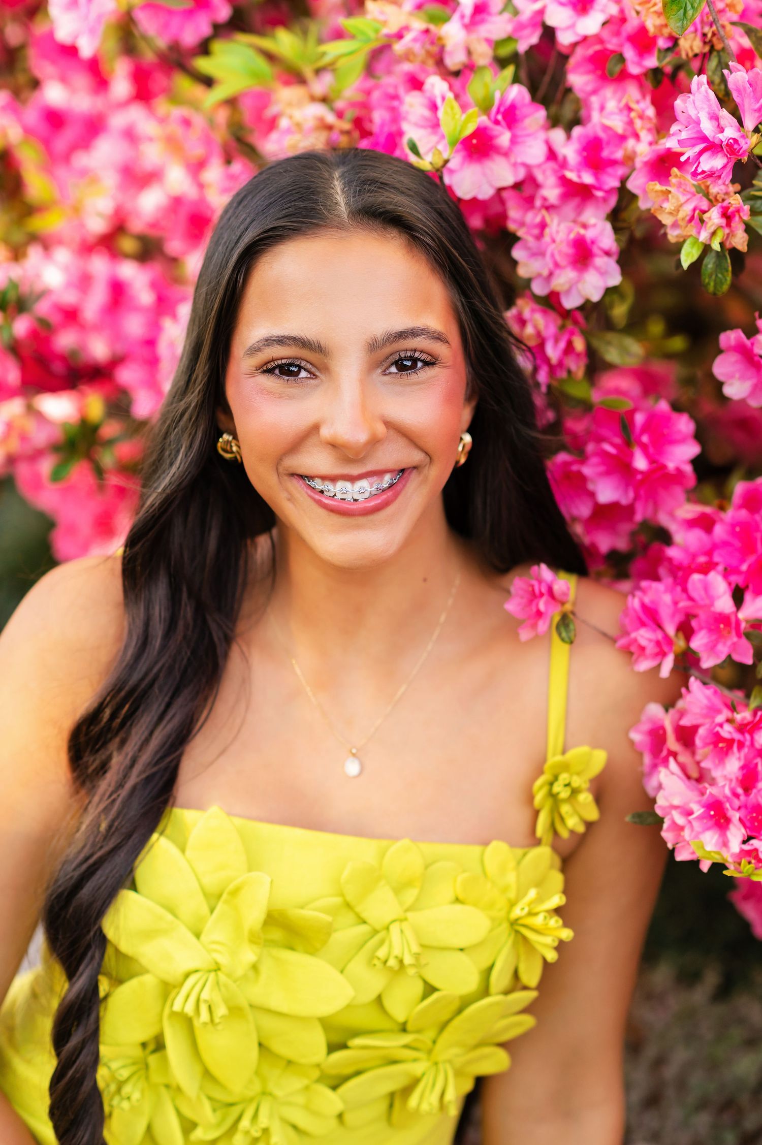 Smiling teenage girl with braces in yellow floral dress posing in front of vibrant pink azalea blooms.