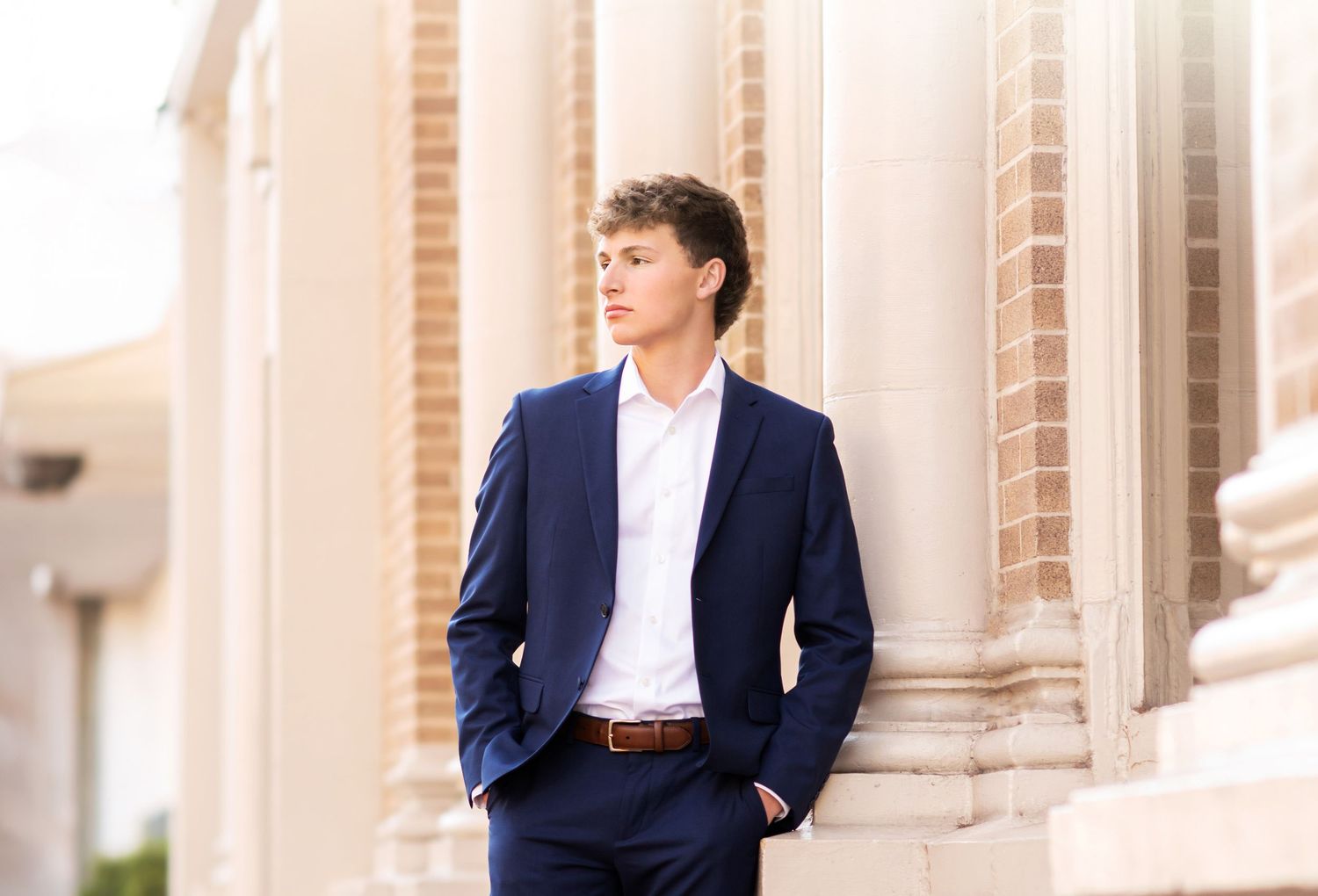 Young man wearing navy suit leaning against stone columns during outdoor senior photo session.