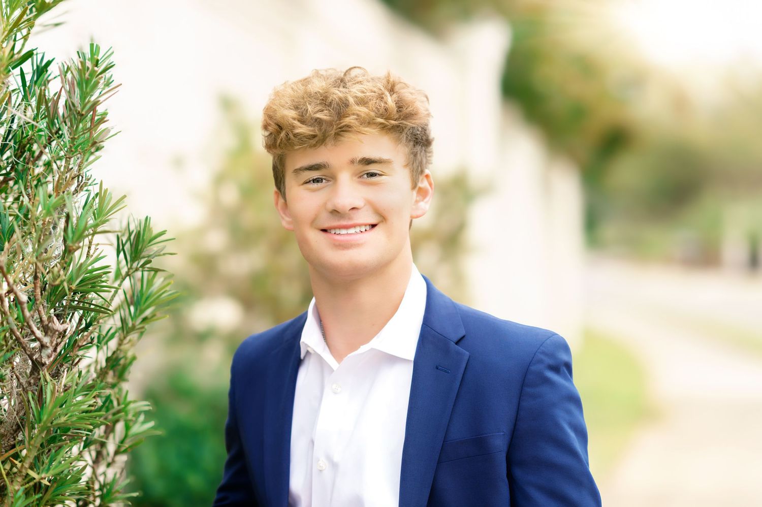 Smiling teenage boy in navy blazer and white shirt poses outdoors near green foliage in soft light.
