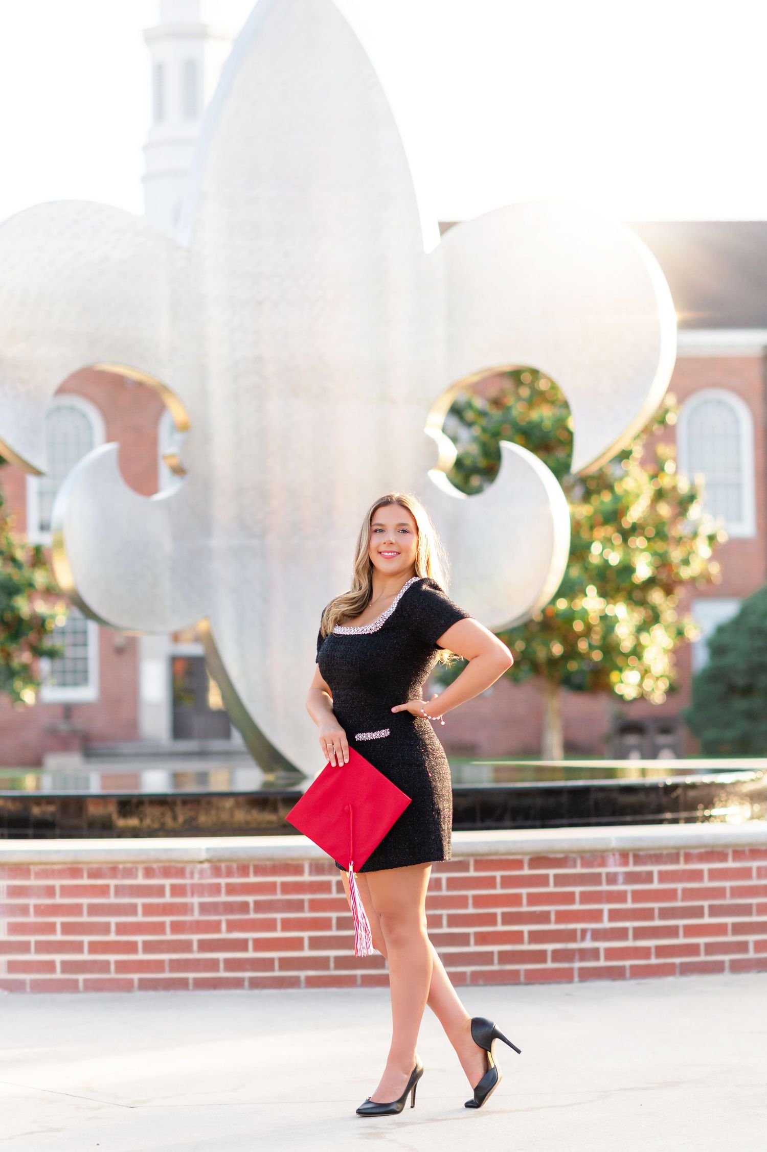Young woman in black dress holds red clutch near large fleur-de-lis sculpture on college campus.