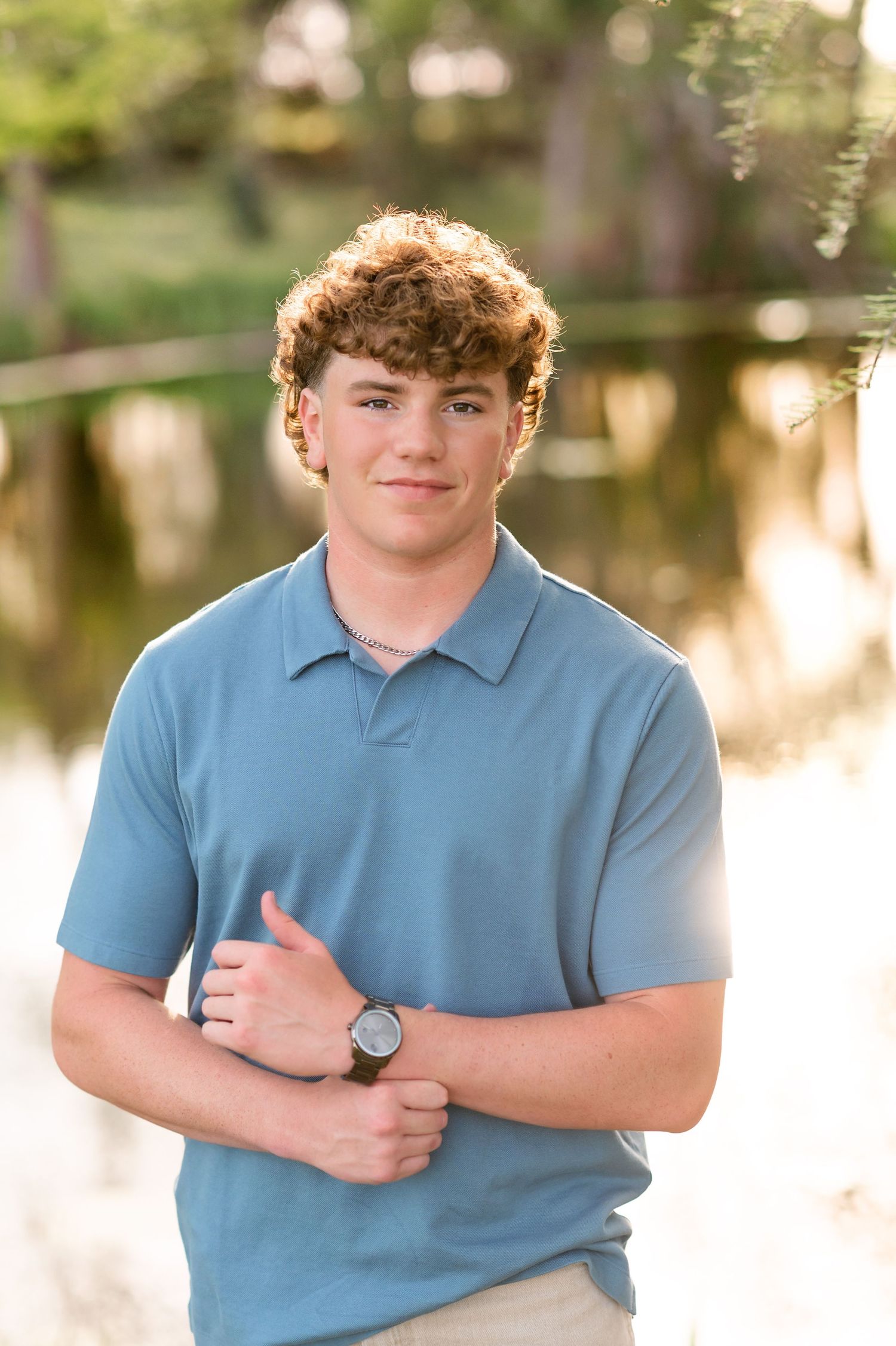 Young man with curly hair wearing a blue polo shirt and watch, giving thumbs up outdoors near a pond.