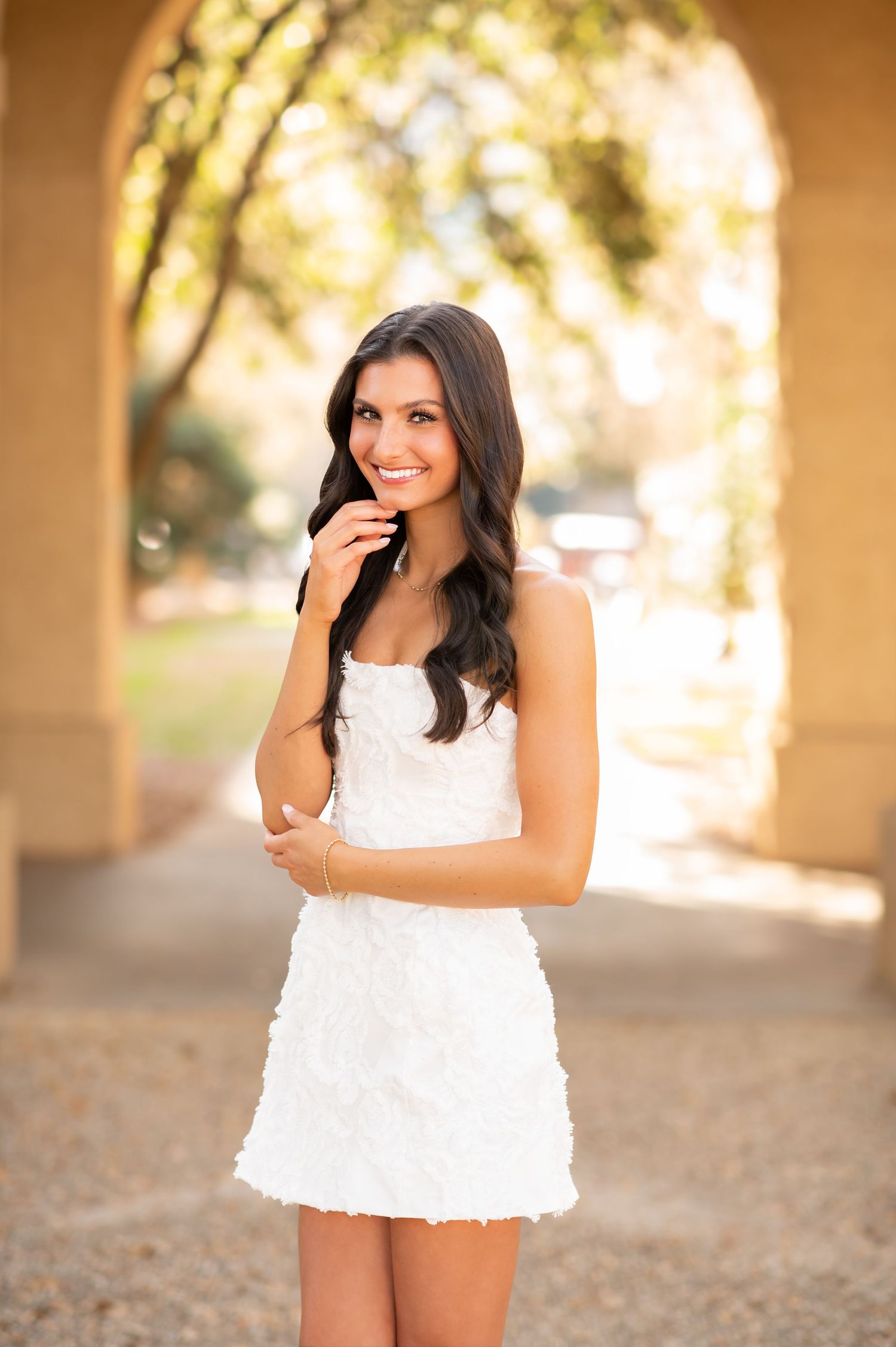 Smiling brunette woman in white mini dress posing under an archway outdoors.