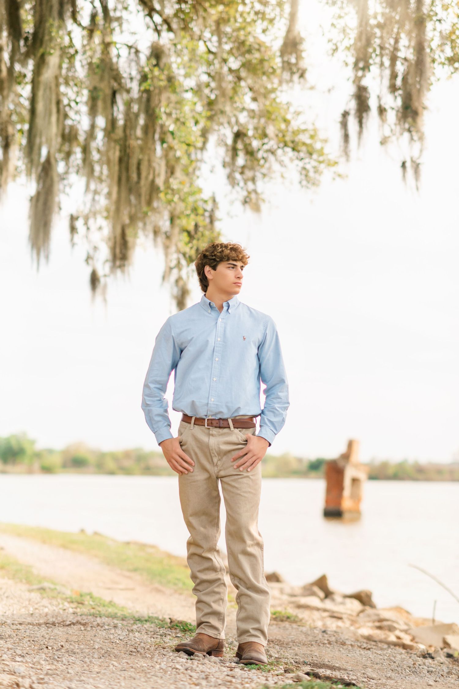 Young man in blue Oxford shirt and khakis stands near a waterway under Spanish moss-draped trees.
