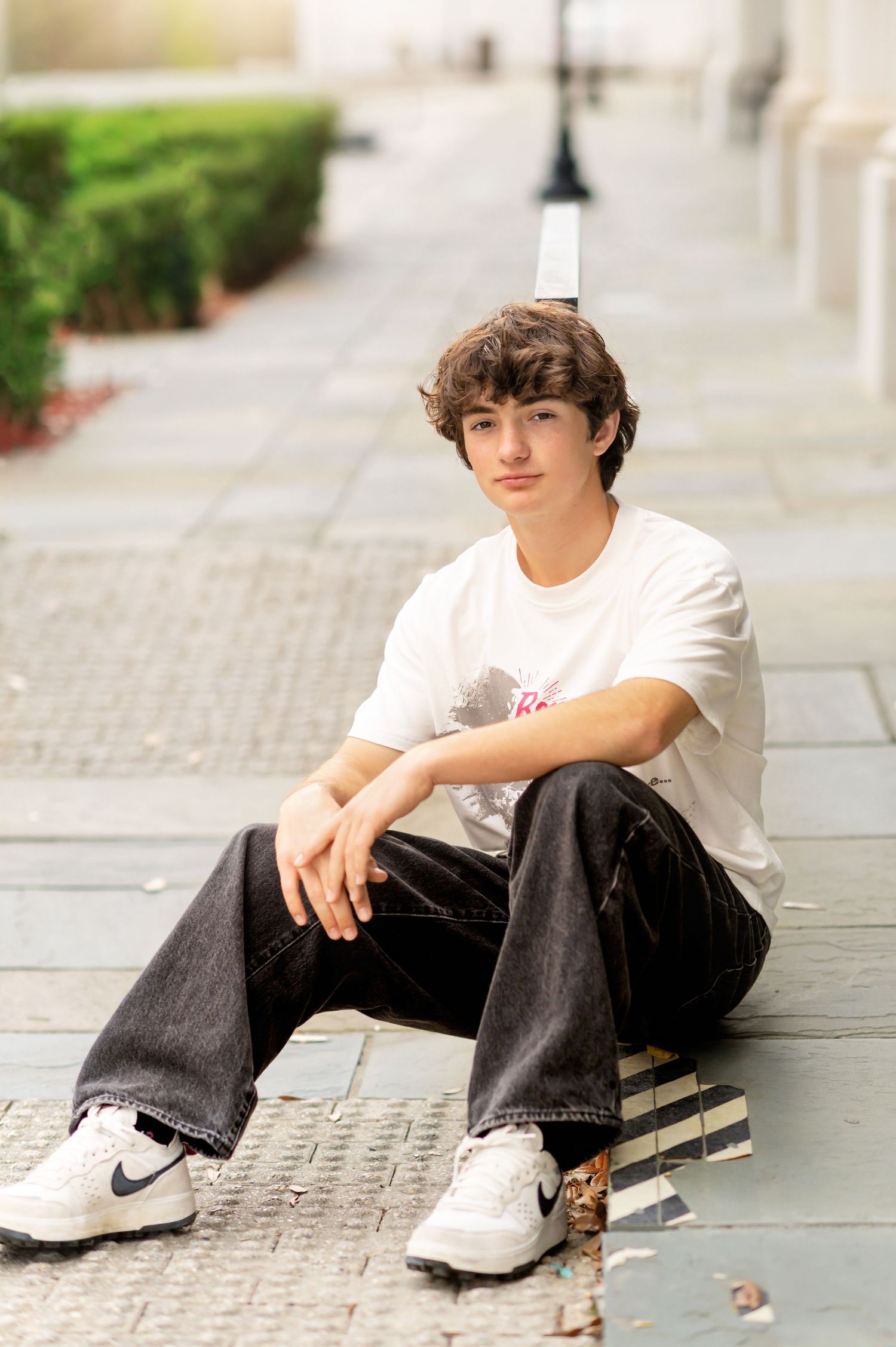 Teen boy in white t-shirt and dark jeans sits relaxed on urban sidewalk wearing Nike sneakers.