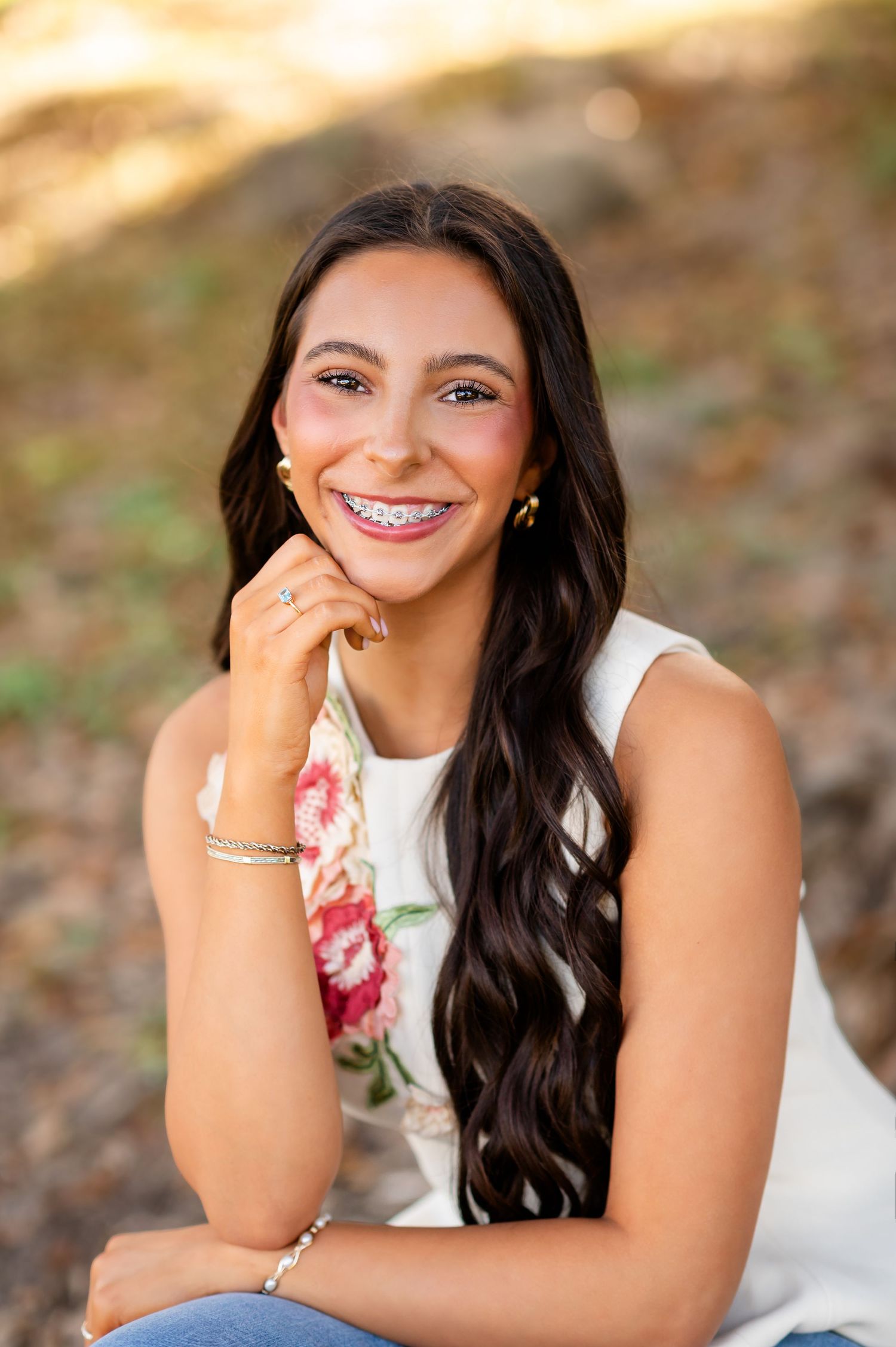 Smiling teenage girl with braces and long dark hair posing outdoors in a white floral top for senior photos.
