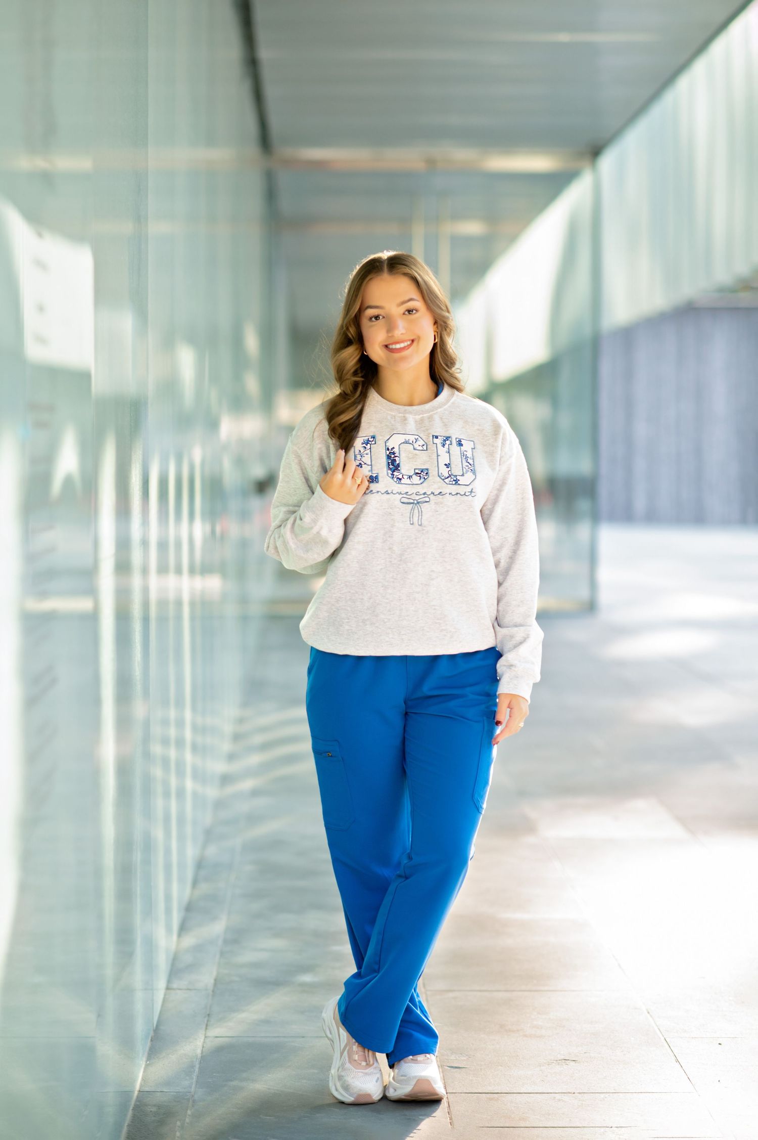 Young woman smiling in gray graphic sweatshirt and blue sweatpants, posing in a modern glass corridor.