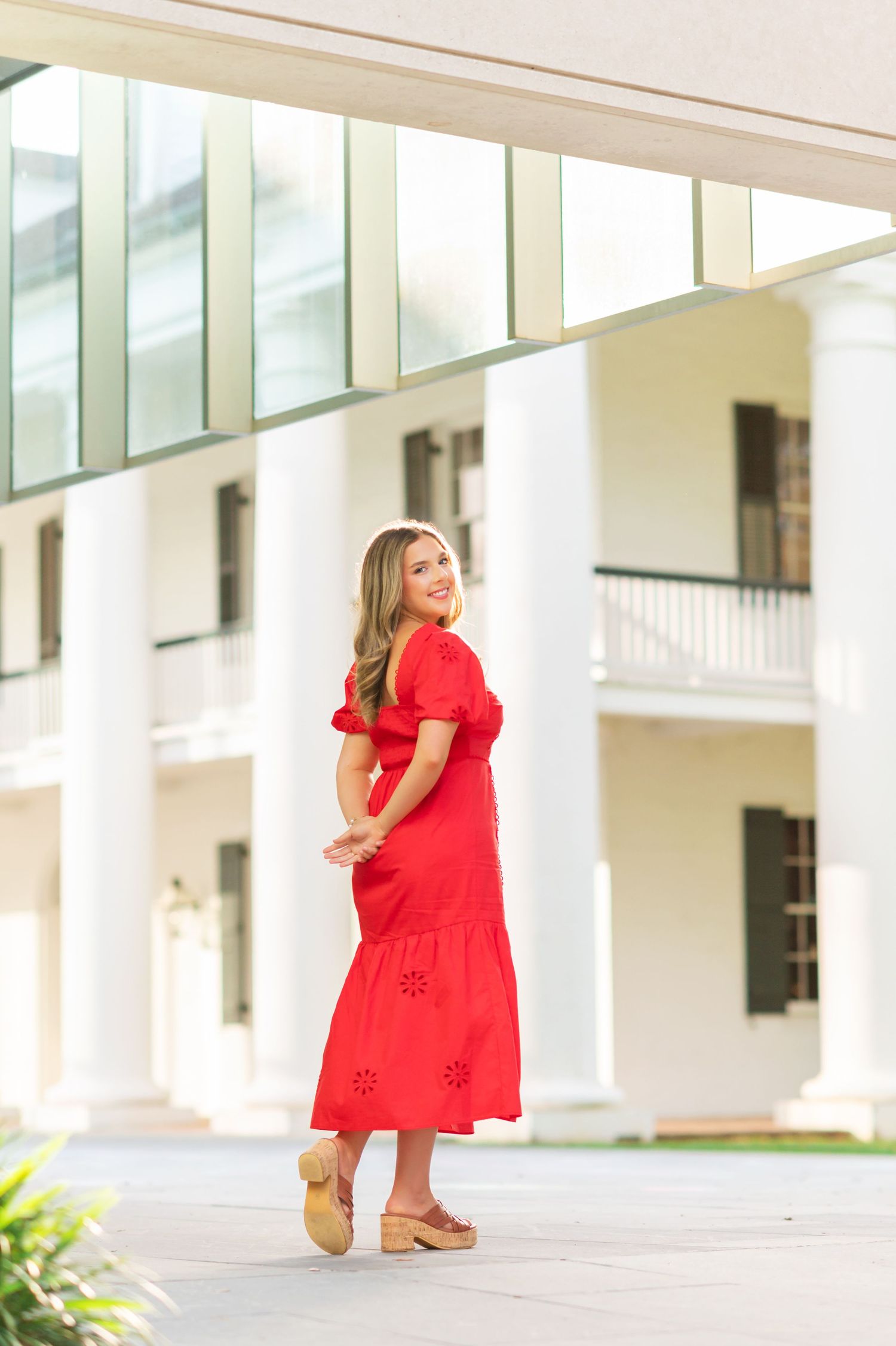 Woman in red midi dress with eyelet details and wedge sandals posing in front of white colonial building.