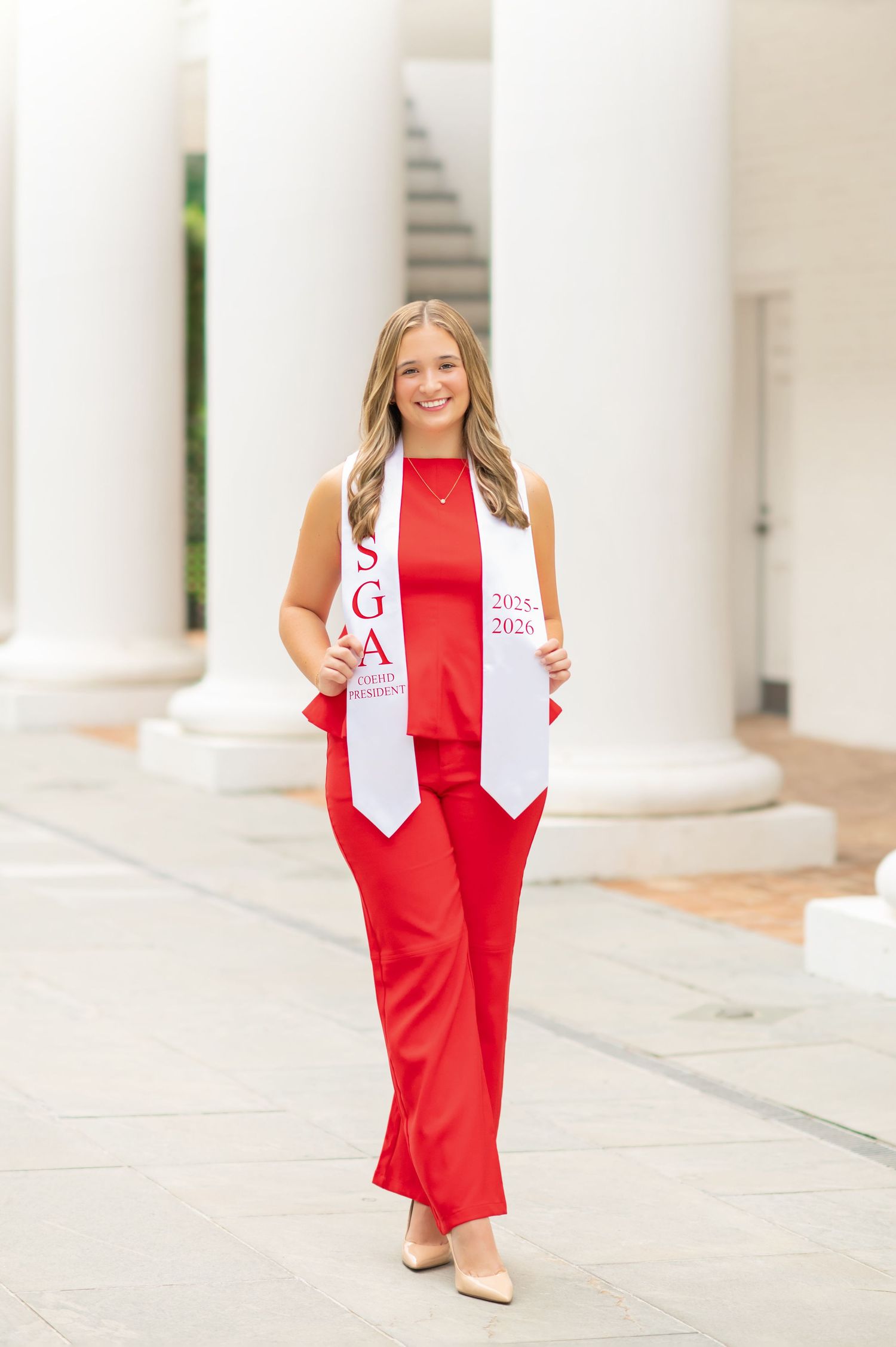 Smiling graduate in red outfit wearing SGA stole poses between white columns for graduation photos.