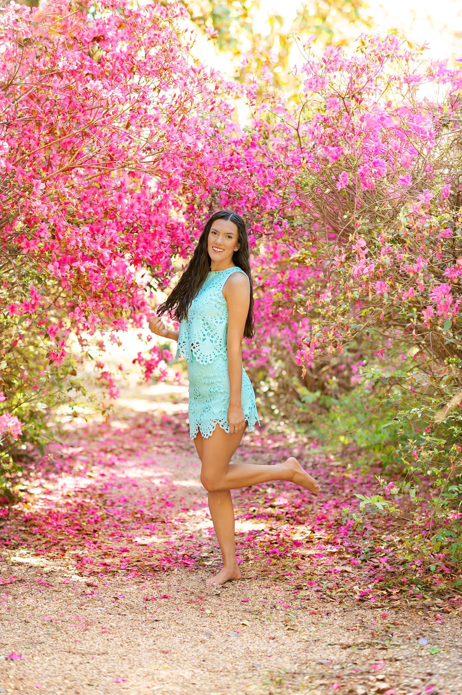Smiling young woman in turquoise lace dress poses playfully among bright pink blooming trees.