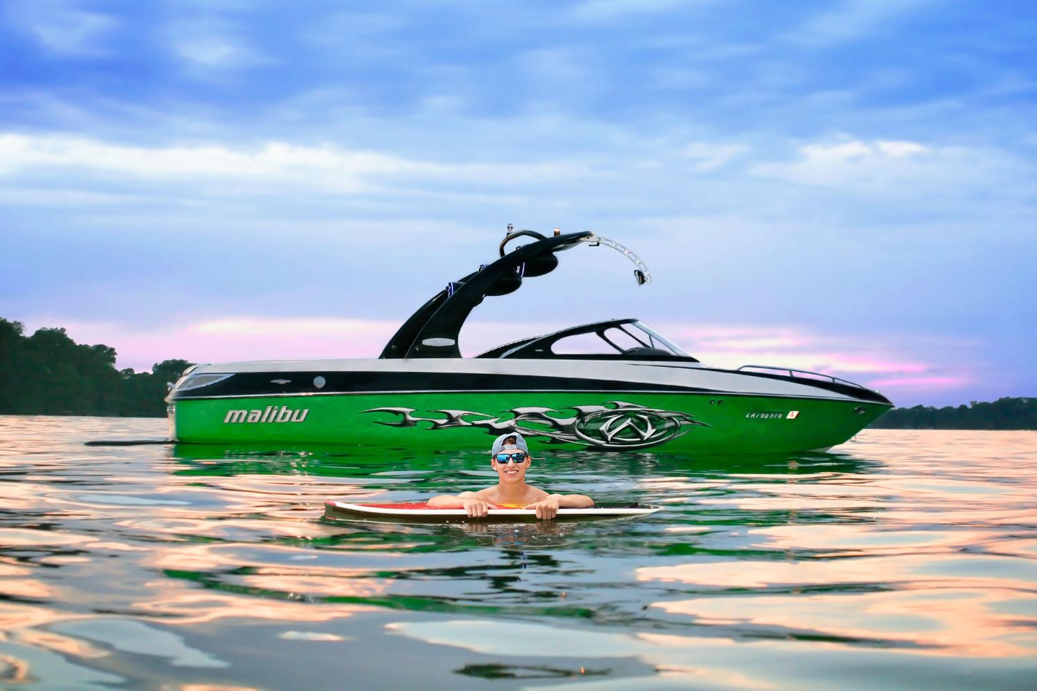 Person floating near green Malibu boat on calm lake water at sunset with colorful sky.