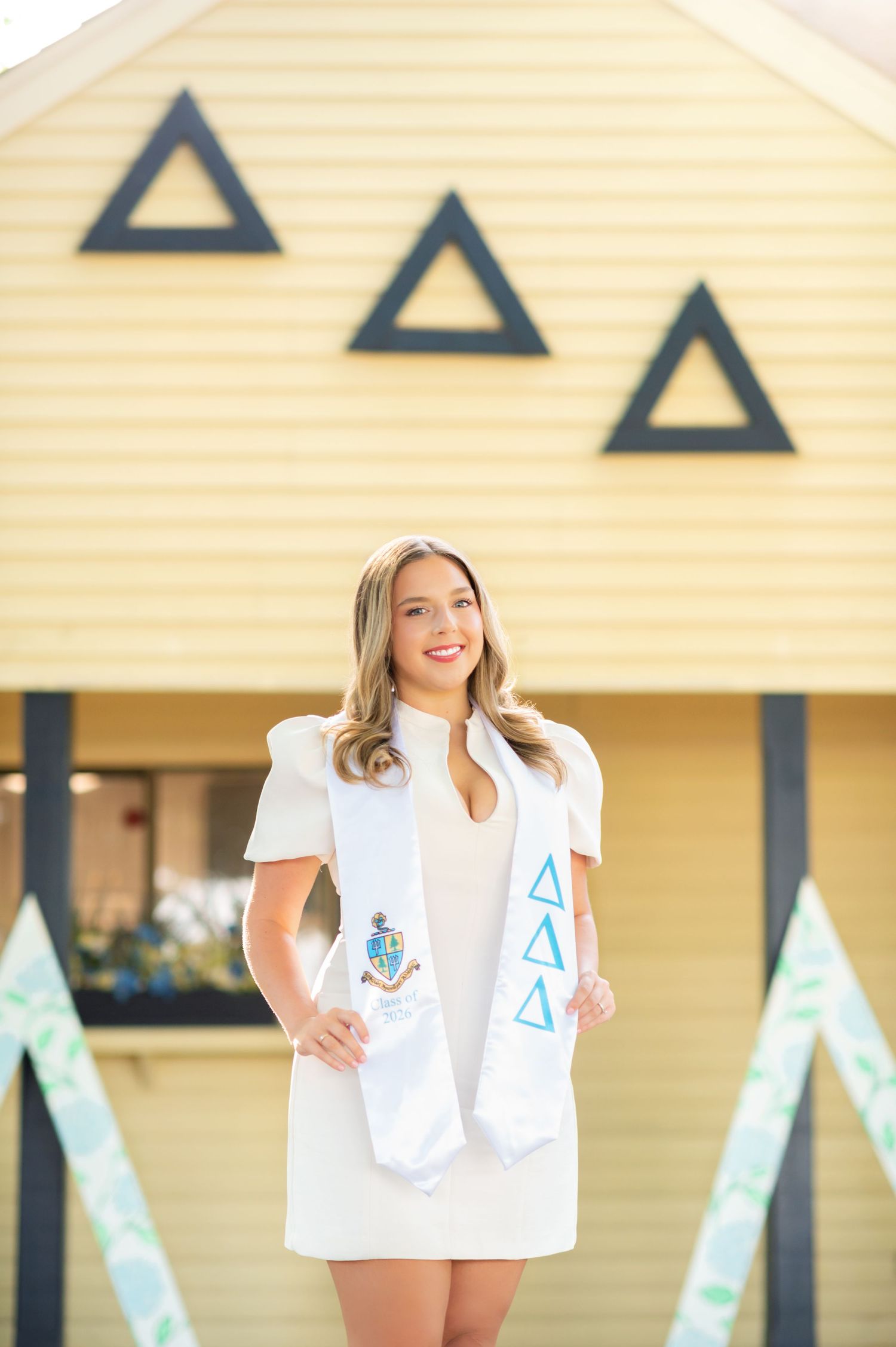 Smiling graduate in white dress wearing Delta Delta Delta sorority stole poses in front of sorority house.