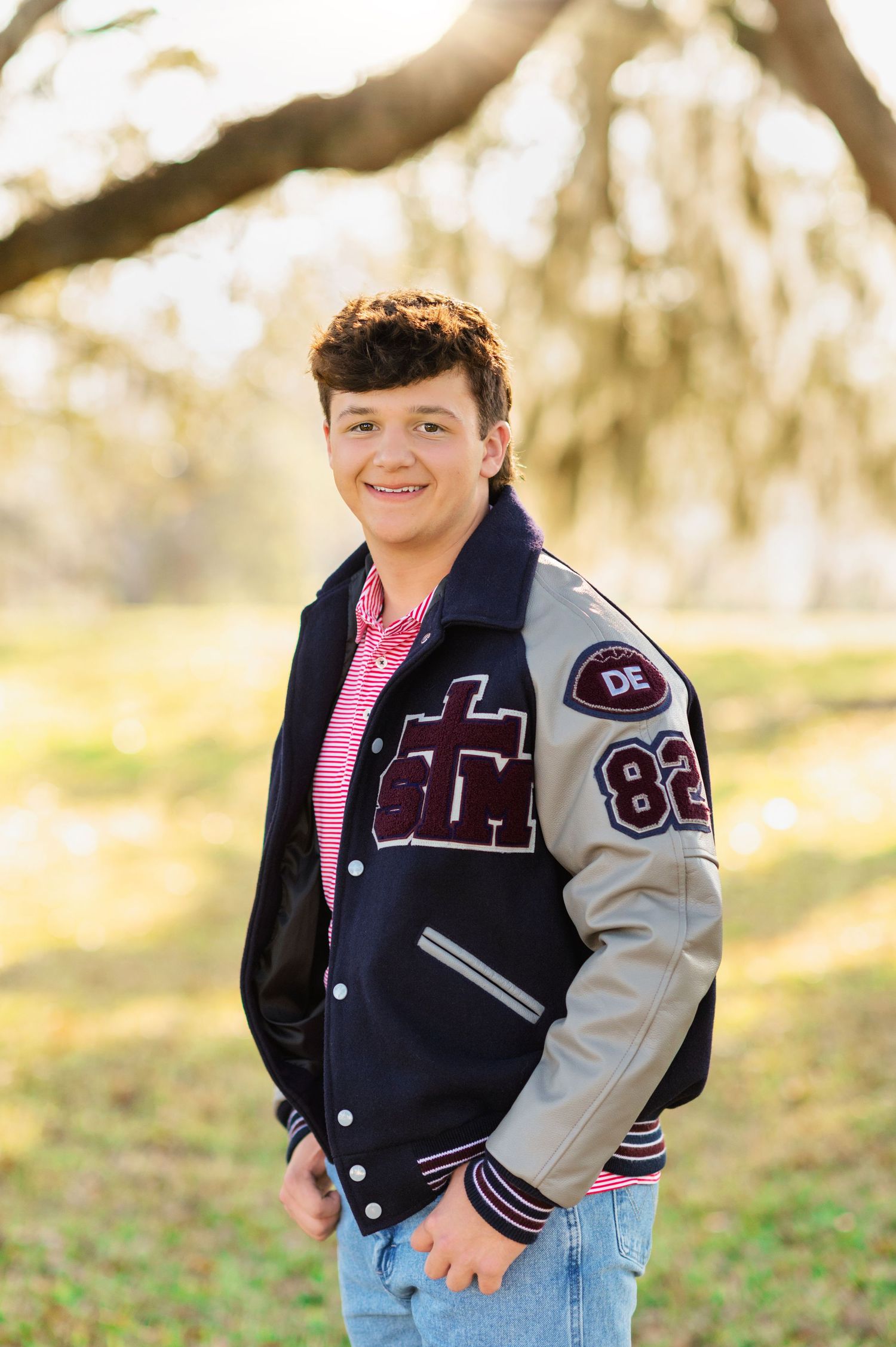 Smiling teen boy wearing a navy letterman jacket with number 82 patch, posed outdoors under a tree in soft golden light.