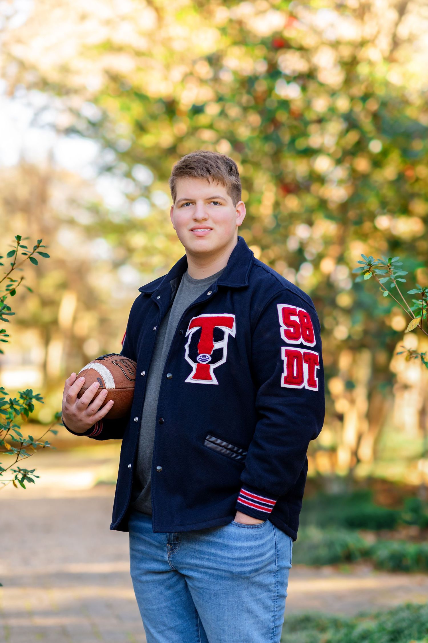 Teen boy in navy letterman jacket holding football poses outdoors among autumn trees for senior portrait photo.