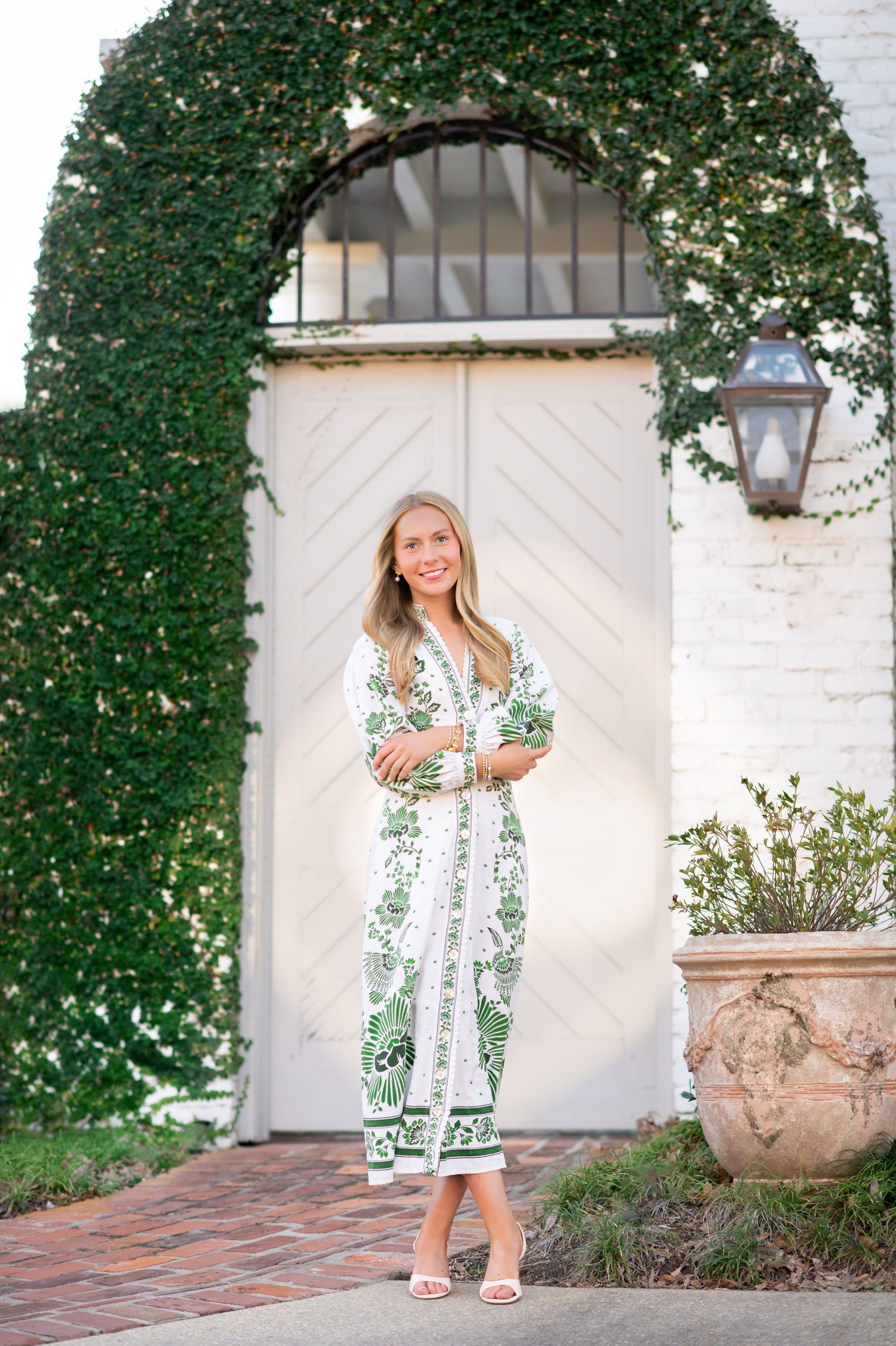 Woman in green and white floral midi dress standing before ivy-covered white arched door with terracotta planter.