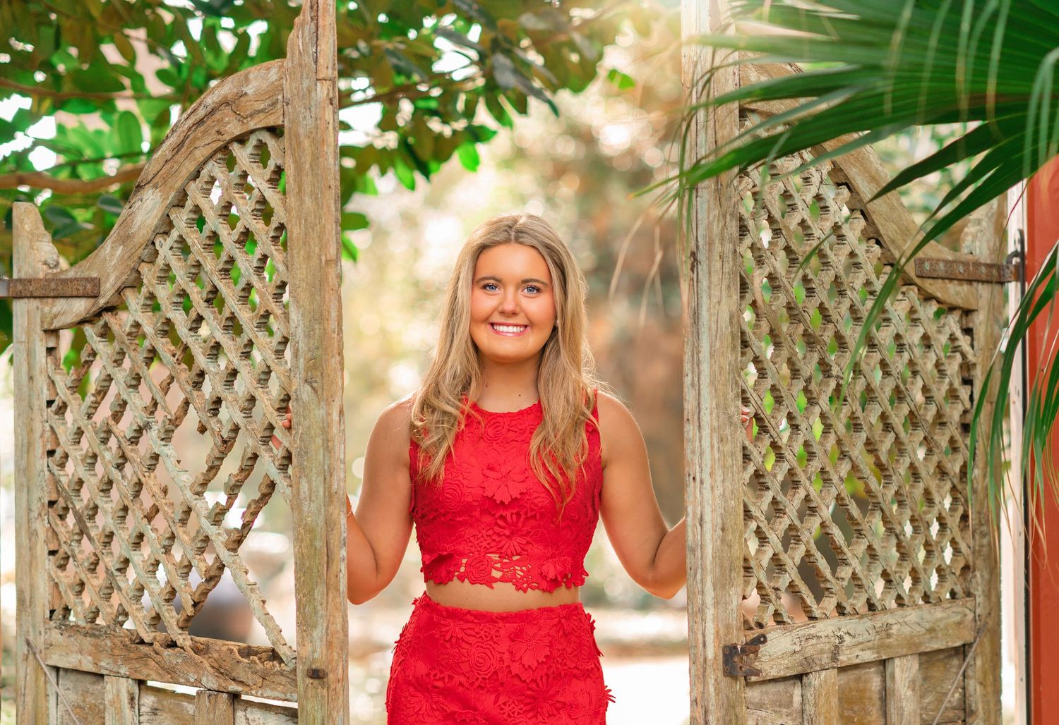 Smiling young woman in red lace two-piece outfit posing between rustic wooden lattice gates with tropical greenery.