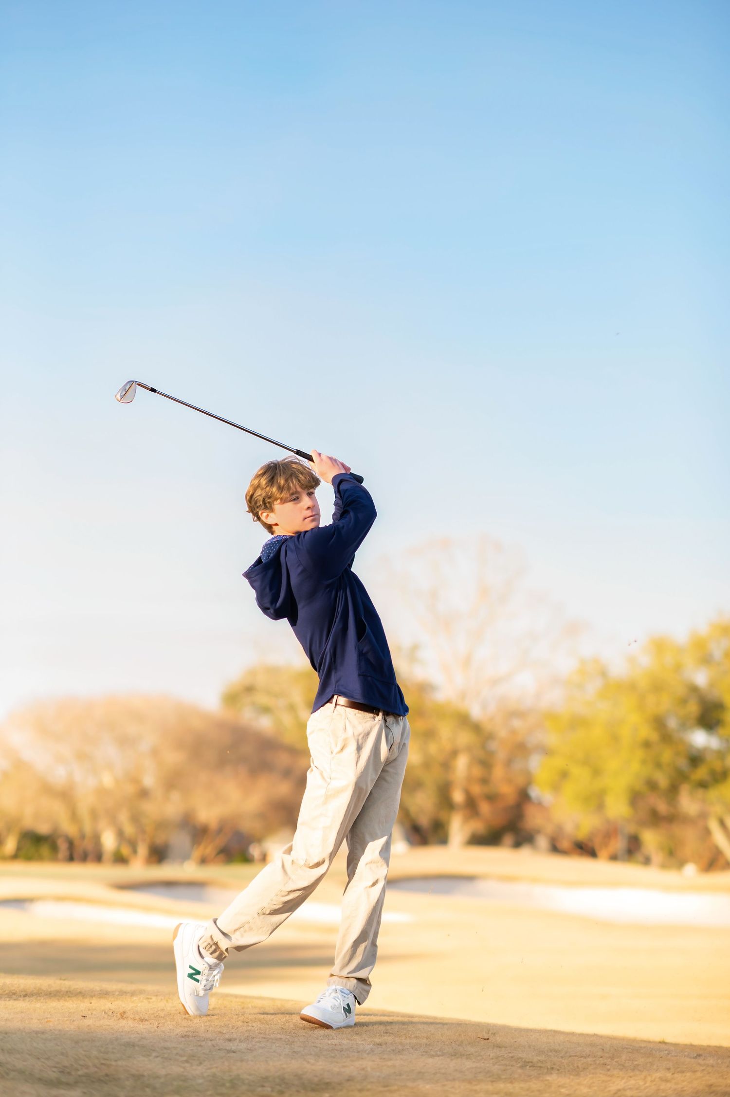 Young golfer completing a full swing on a sunny golf course, wearing a navy sweater and khaki pants.
