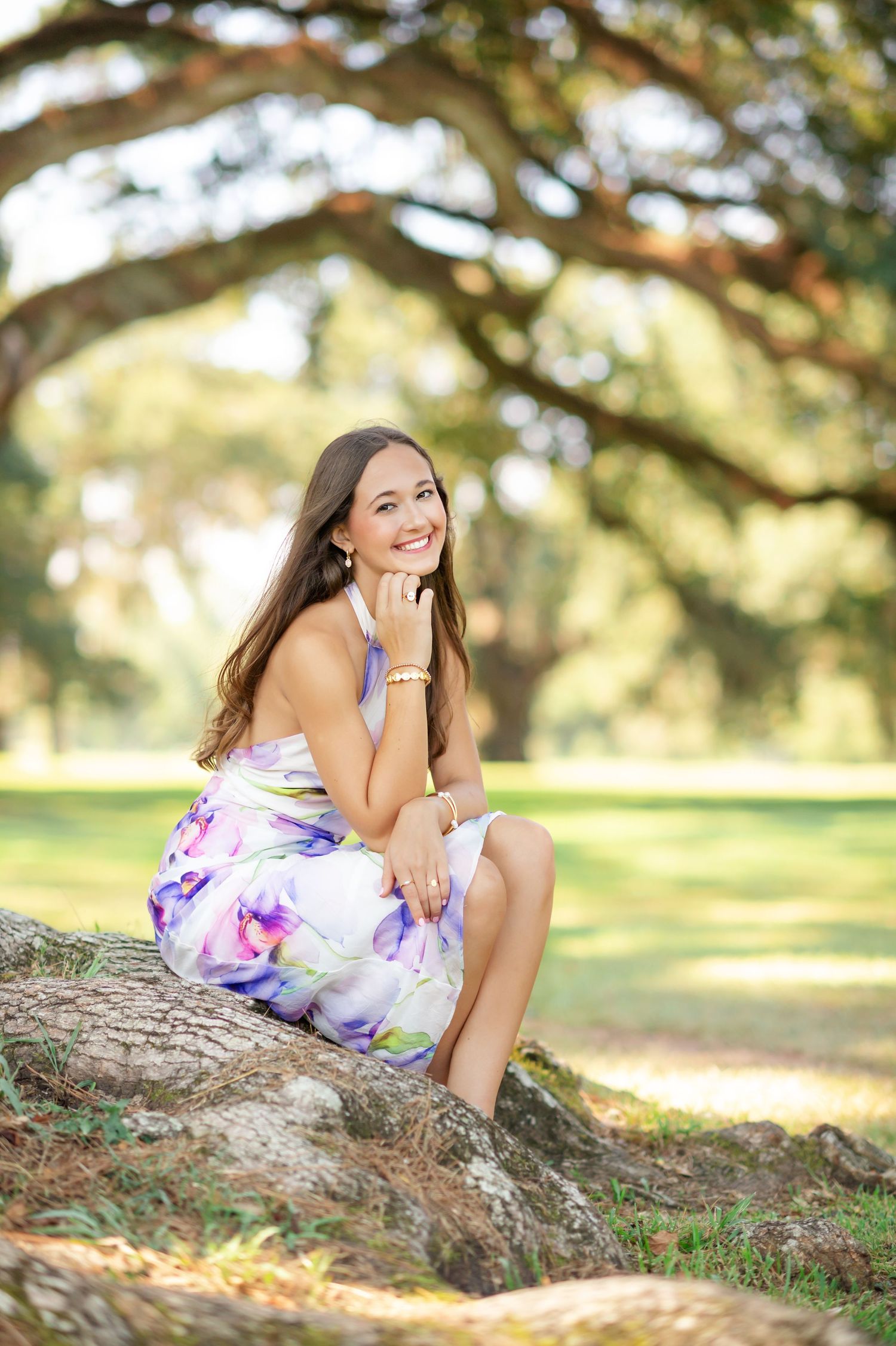 Young woman in floral dress sitting on tree roots in a sunlit park, smiling gracefully.