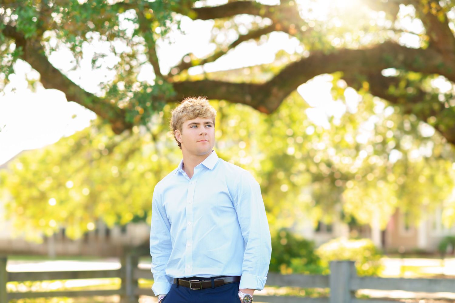 Young man in light blue dress shirt standing under a large oak tree near a wooden fence.