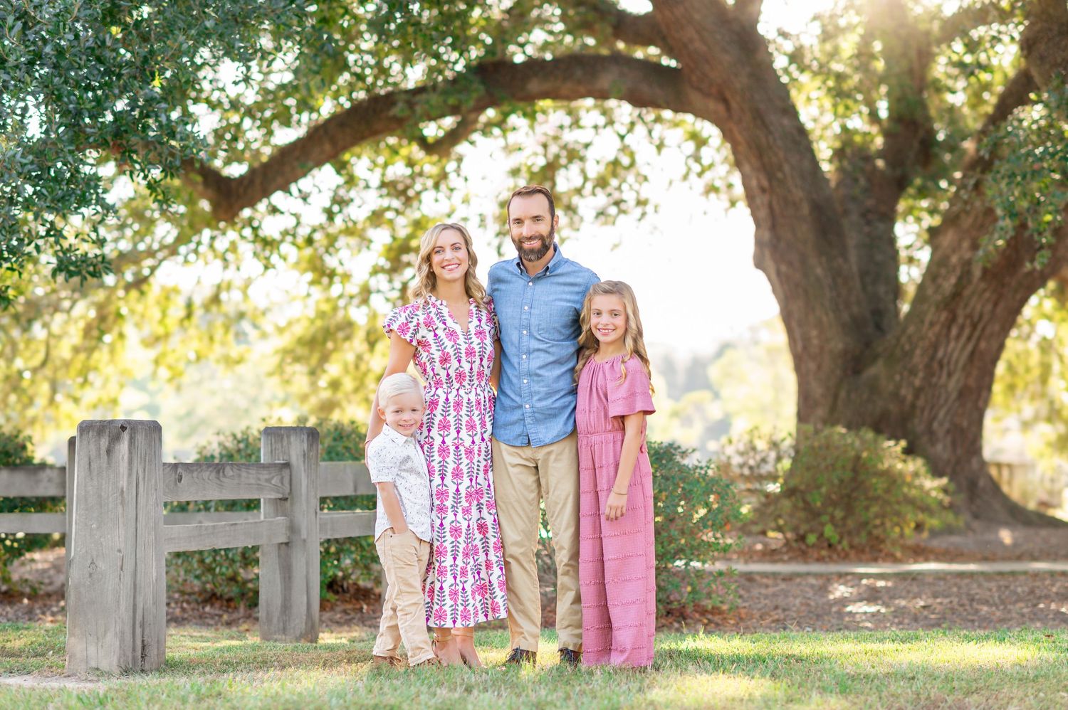 Family of four standing together under a large oak tree in a sunny park in coordinating pink and blue outfits.
