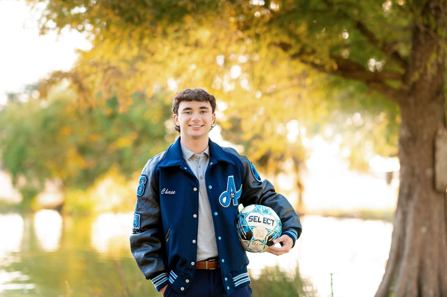 Teen boy in blue letterman jacket holding a soccer ball outdoors near a pond with autumn trees.