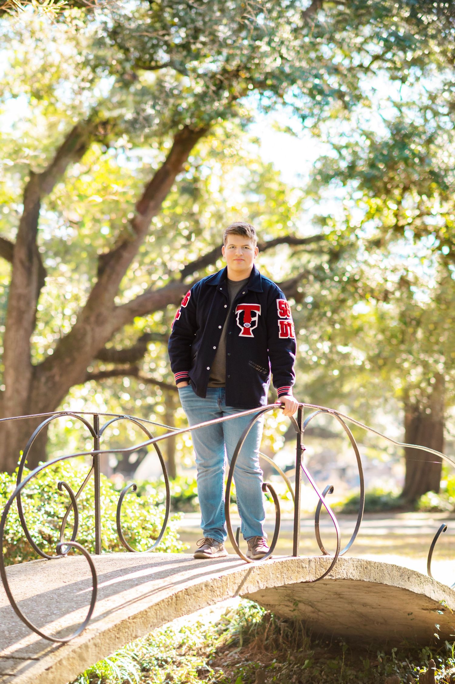 Teen boy in varsity jacket poses on ornate bridge surrounded by sunlit trees in a park setting.