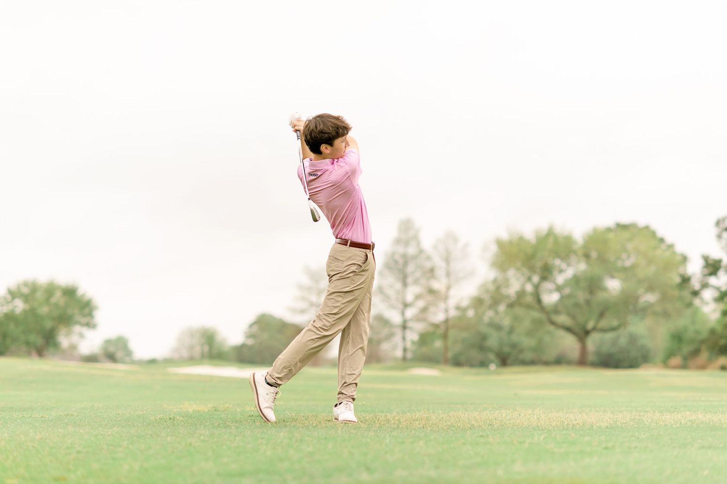 Golfer in pink shirt completing a full swing on a lush green golf course.