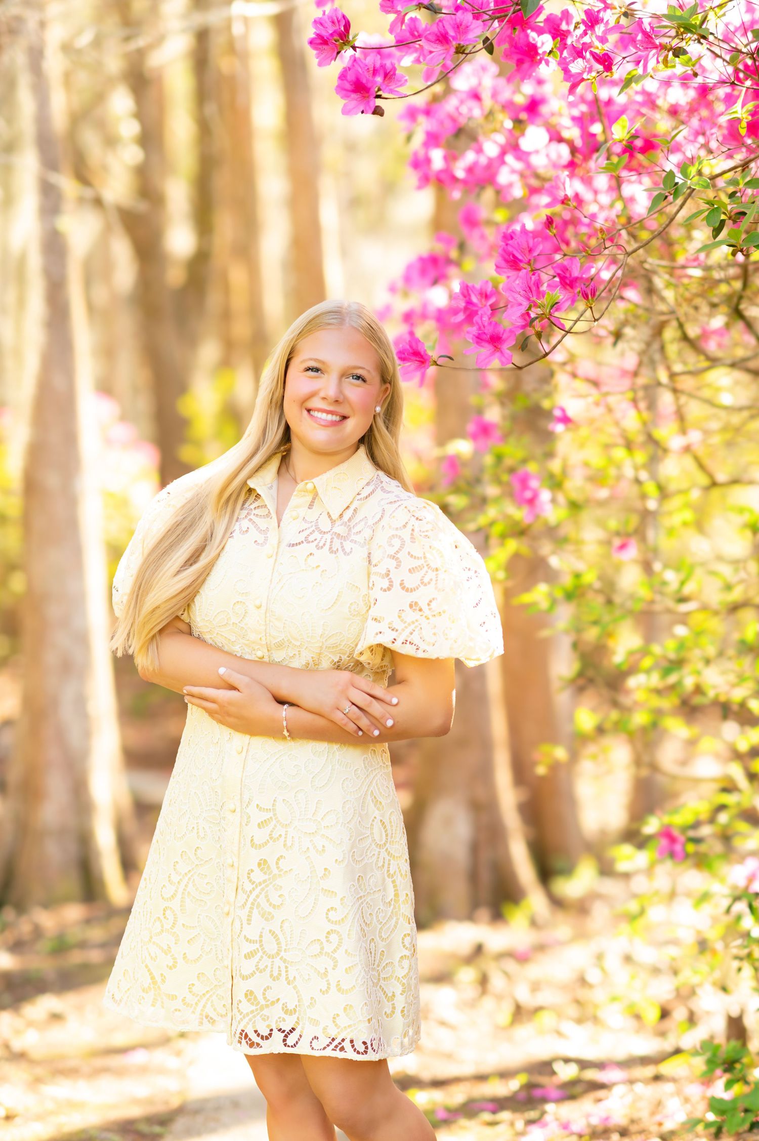 Smiling blonde woman in cream lace dress stands beside vibrant pink bougainvillea in sunny park.