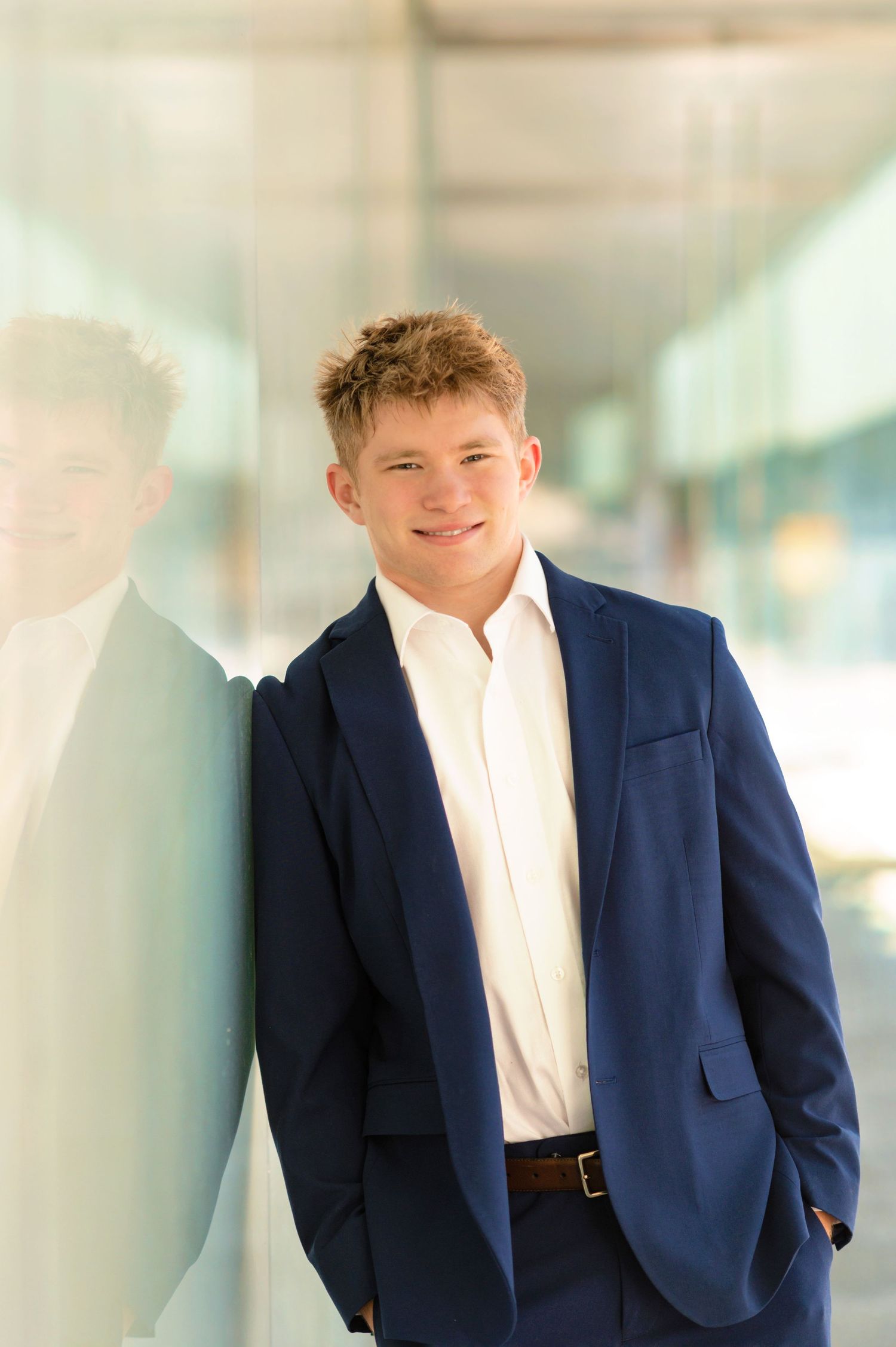 Young man in navy blue suit and white shirt smiling confidently, leaning against a glass wall outdoors.