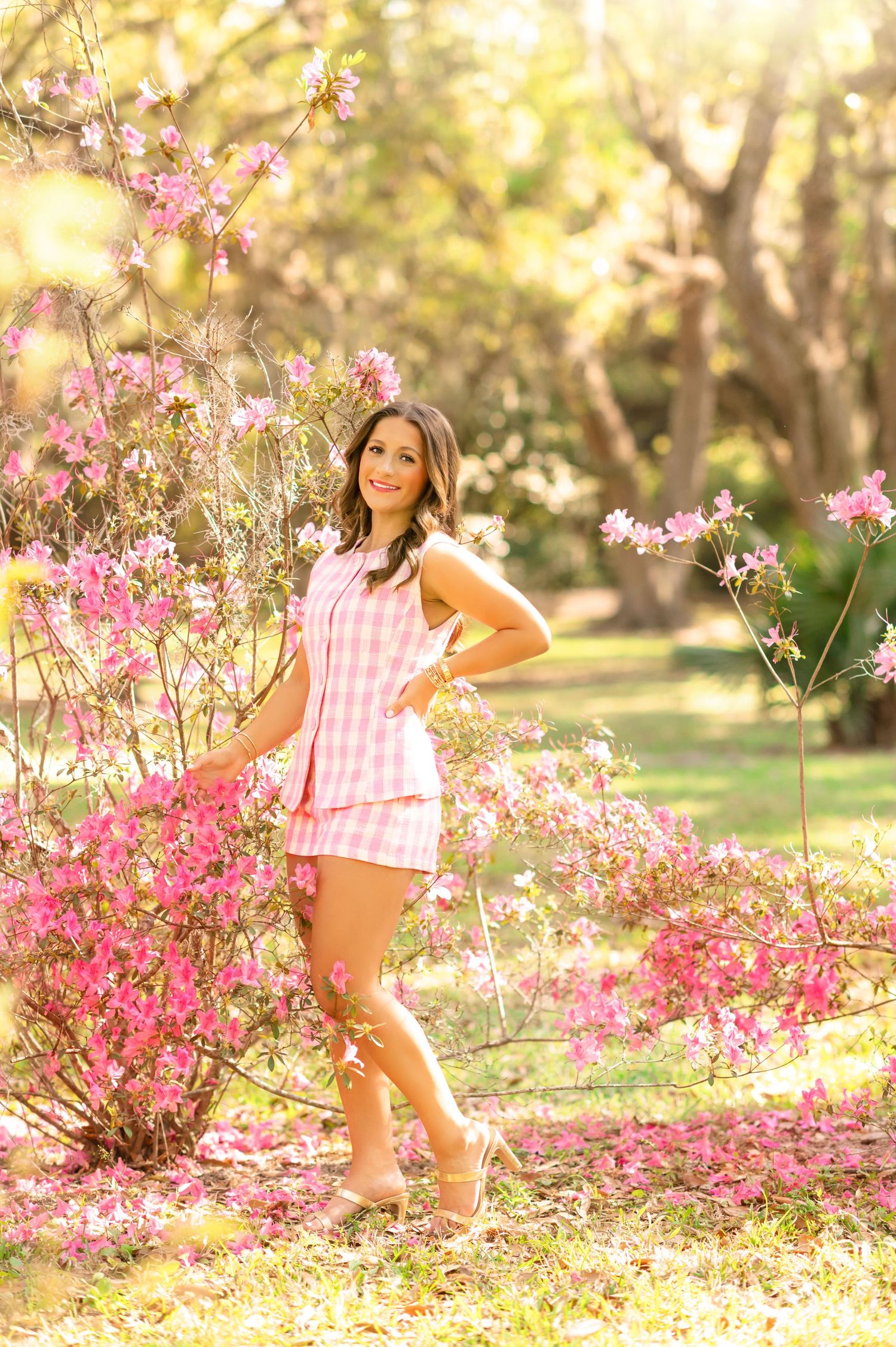 Young woman in pink gingham outfit posing among blooming pink azalea bushes in a sunlit garden.