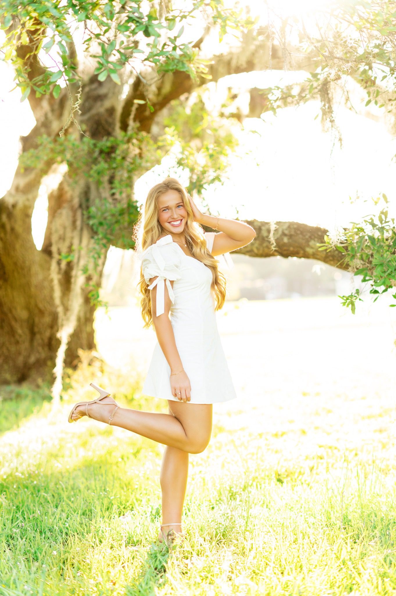Smiling blonde woman in white dress posing under sunlit oak tree with one leg raised.