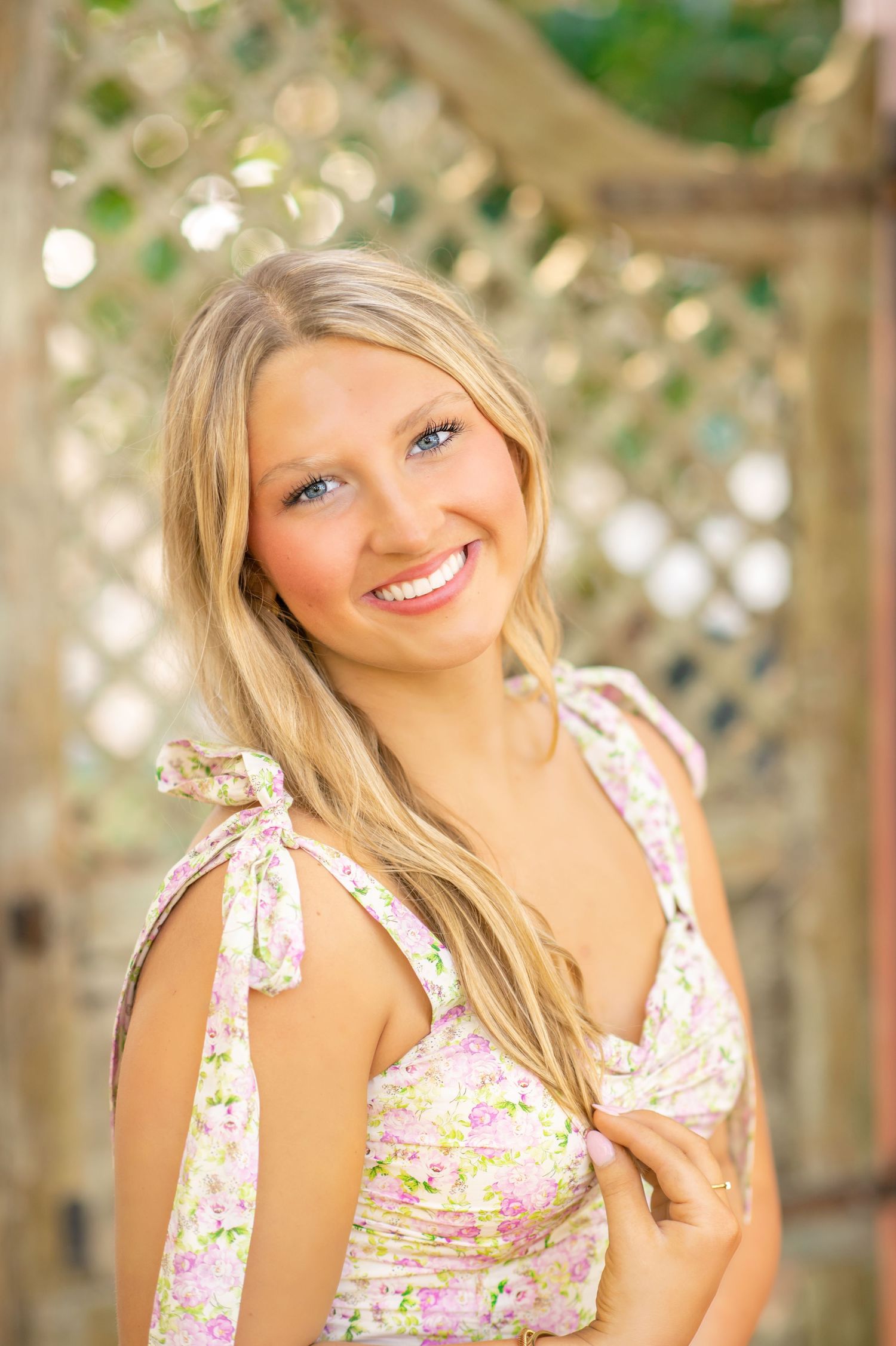 Smiling blonde woman with blue eyes wears floral sundress near a wooden lattice garden structure.