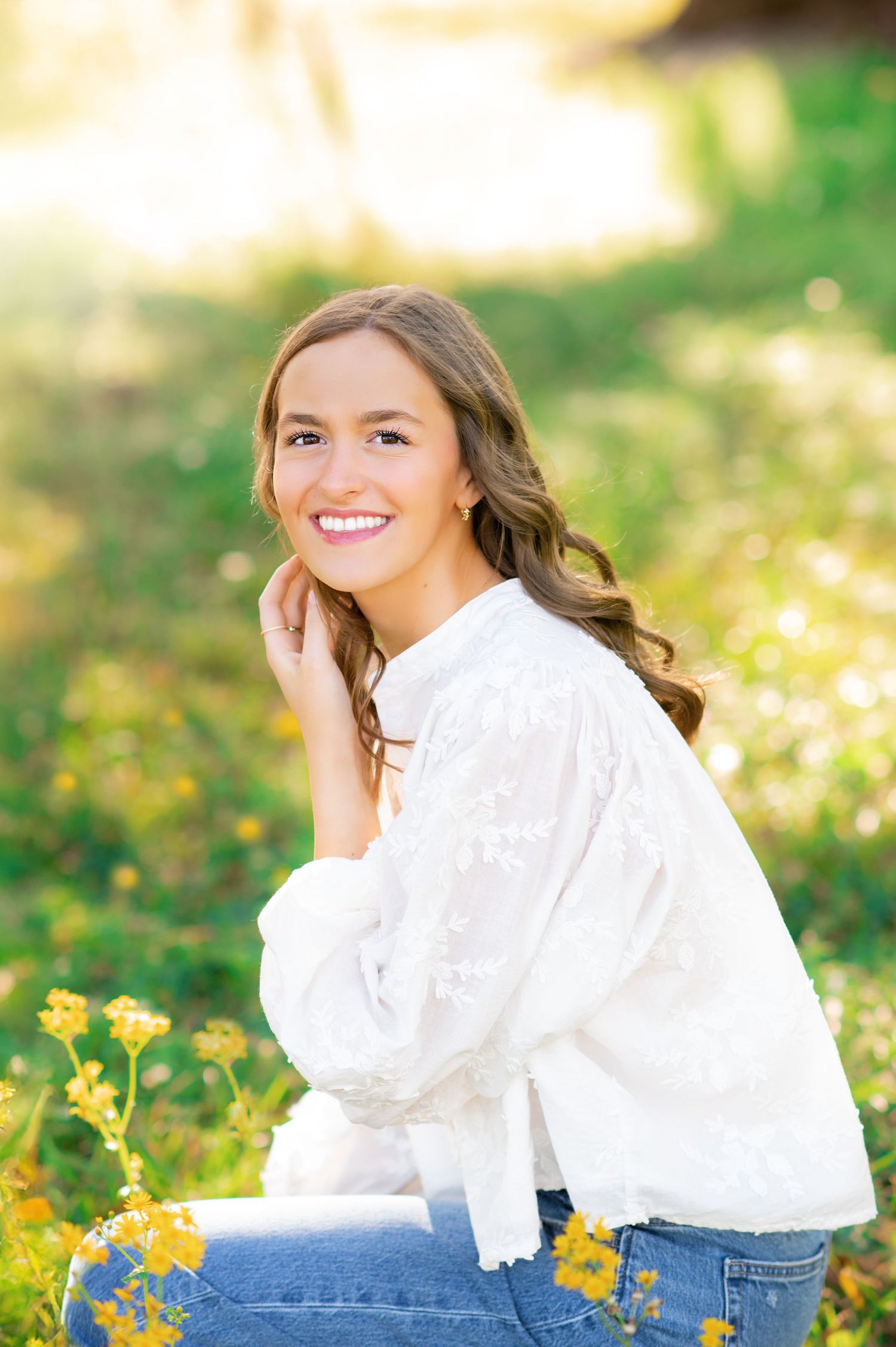 Smiling young woman in white embroidered blouse sitting among yellow wildflowers in sunny green field.